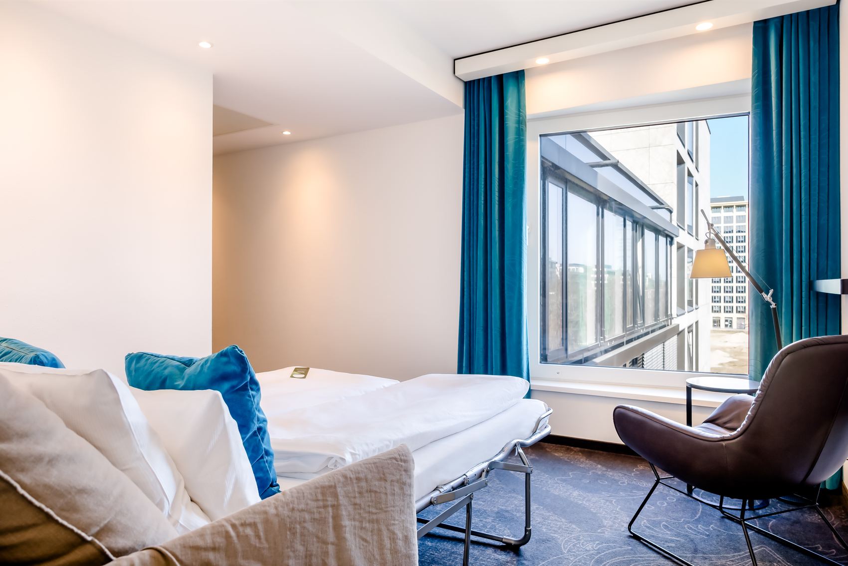 Hell eingerichtetes Hotelzimmer mit gemütlichem Bett, blauen Vorhängen und einem stilvollen Sessel am Fenster mit Stadtblick. Ruhige, moderne Atmosphäre ideal zum Entspannen oder Arbeiten.