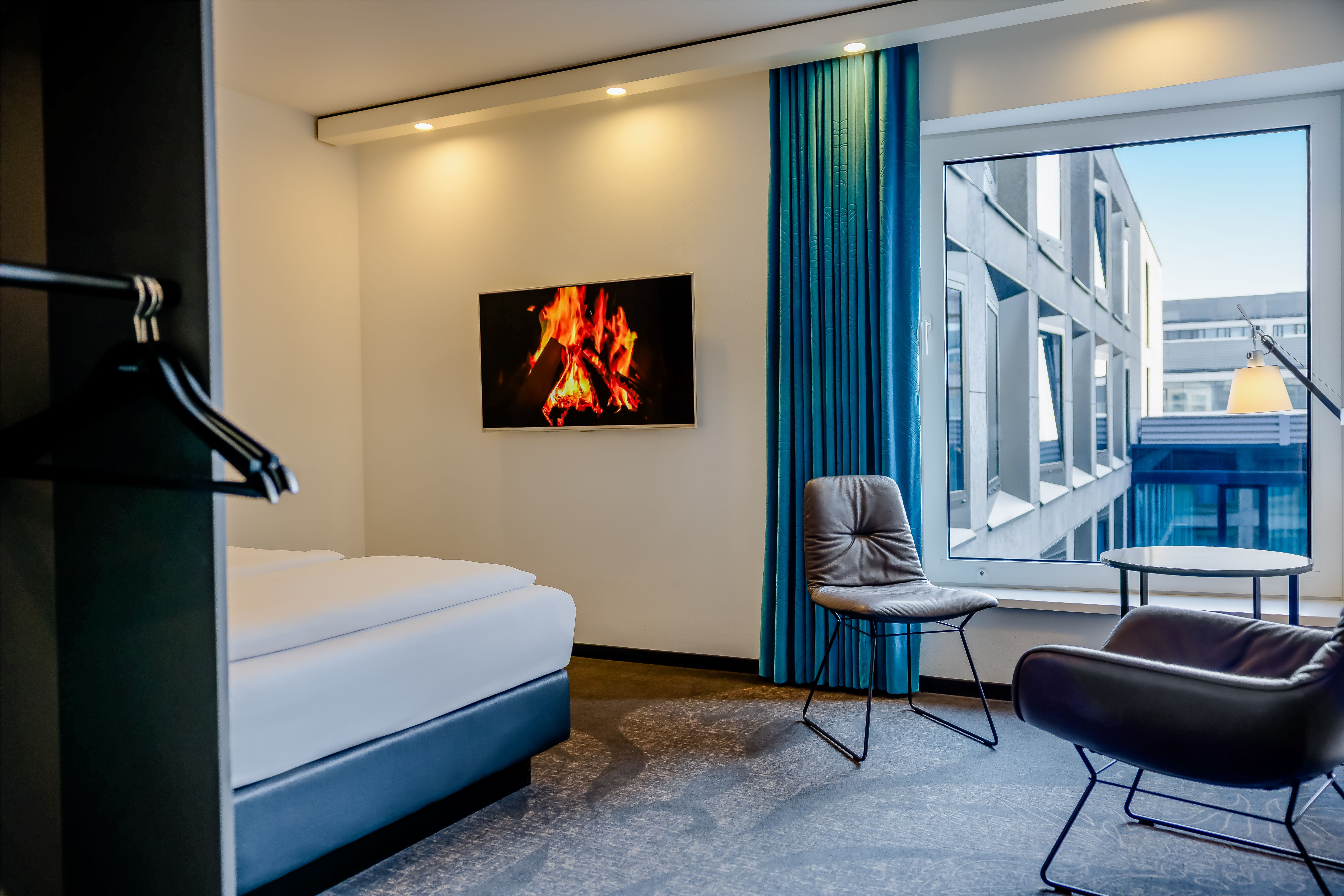 Modernes Hotelzimmer mit gemütlichem Bett, zwei bequemen Sesseln und großem Fenster mit Blick auf benachbarte Gebäude. An der Wand hängt ein Bildschirm, der ein prasselndes Kaminfeuer zeigt, was eine warme und einladende Atmosphäre schafft.