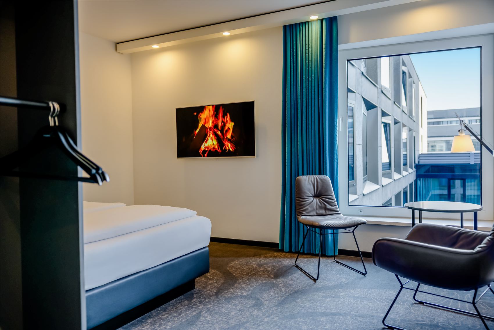Modernes Hotelzimmer mit gemütlichem Bett, zwei bequemen Sesseln und großem Fenster mit Blick auf benachbarte Gebäude. An der Wand hängt ein Bildschirm, der ein prasselndes Kaminfeuer zeigt, was eine warme und einladende Atmosphäre schafft.