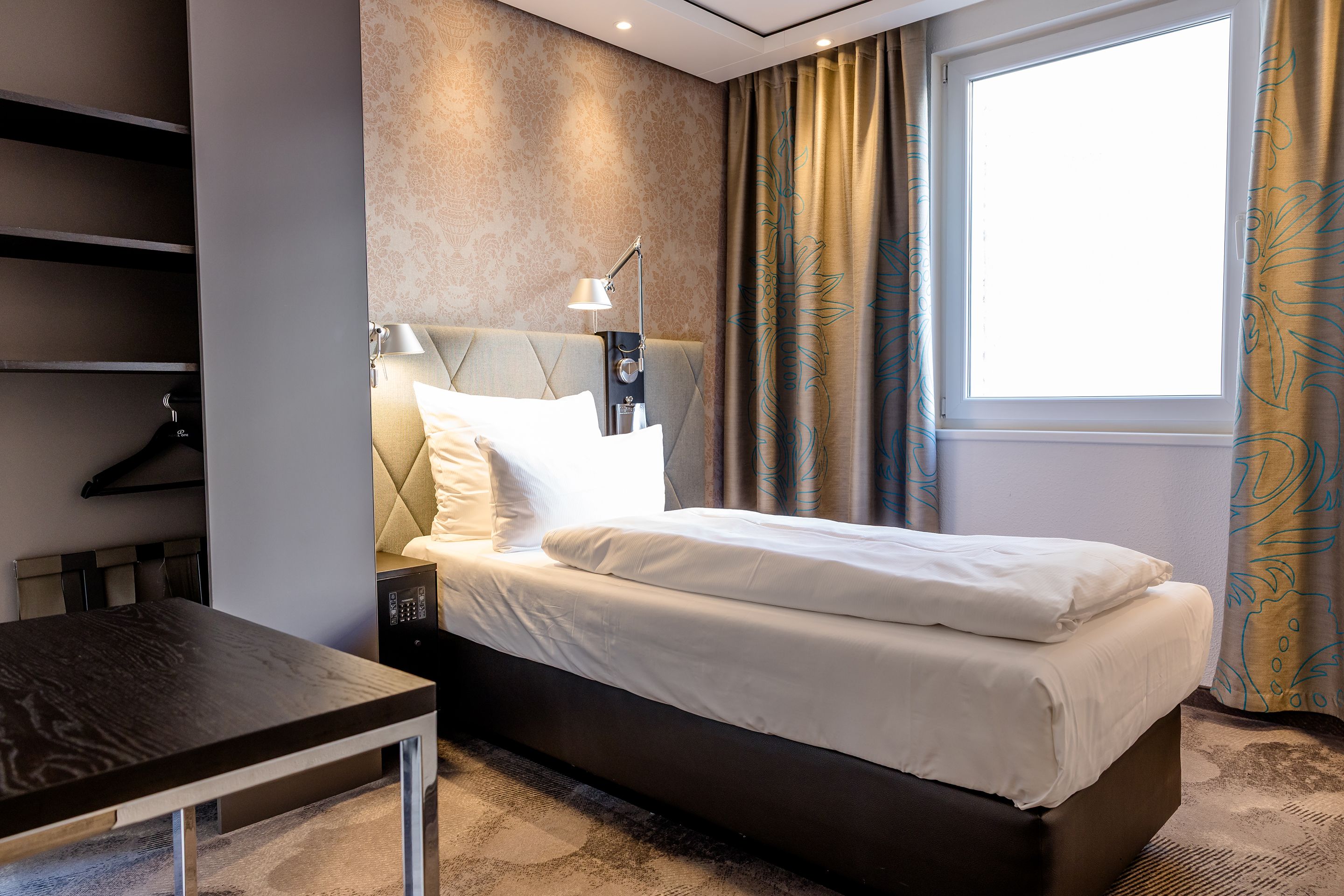 Hotel Motel One München-Sendlinger Tor Helles Einzelzimmer mit modernem Design, komfortablem Bett, weichen Kissen, Leselampen und einem Fenster mit Blick nach draußen. Vorhänge und dezente Wandgestaltung erzeugen eine gemütliche Atmosphäre.