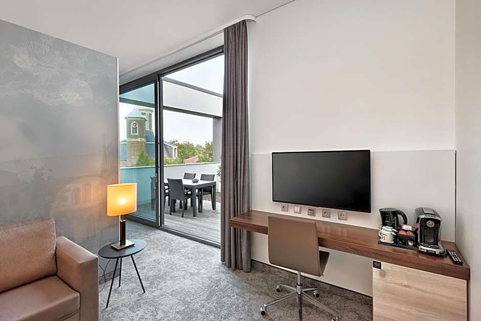 Helles Hotelzimmer mit gemütlichem Sessel, Schreibtisch und großer Wandhalterung für Fernseher. Große Glas-Schiebetür ermöglicht Zugang zu Balkon mit Sitzbereich und Blick ins Grüne. Raum wirkt modern und einladend.