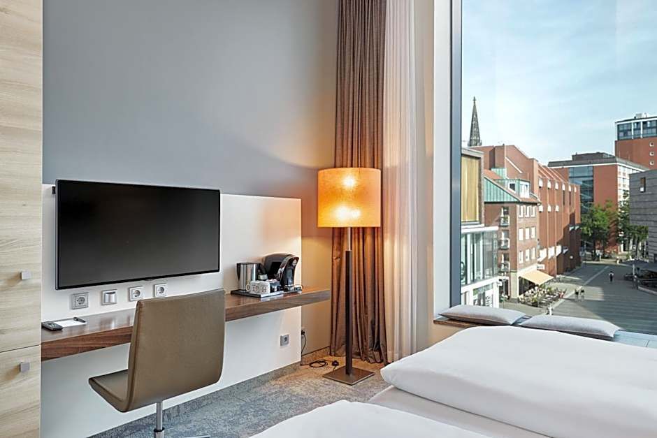 Modernes Hotelzimmer mit Doppelbett, großem Fenster mit Stadtblick, Schreibtisch mit Stuhl, Flachbildfernseher und Stehlampe für eine angenehme Arbeits- und Entspannungsatmosphäre.