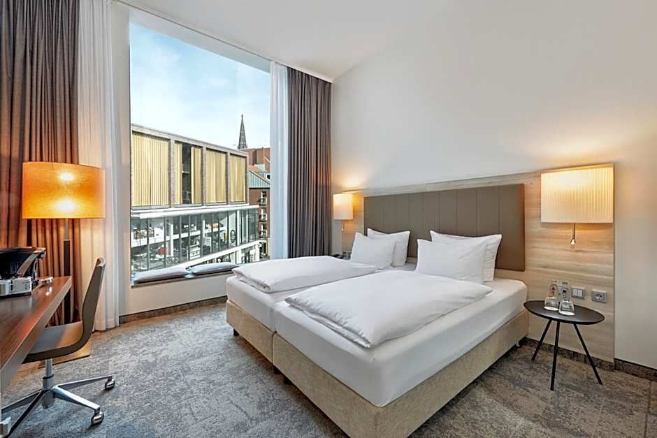Hell eingerichtetes Hotelzimmer mit großem Doppelbett, modernem Schreibtisch, bodentiefem Fenster und Blick auf eine städtische Umgebung. Gemütliche Atmosphäre mit warmem Licht und klarer, aufgeräumter Gestaltung.