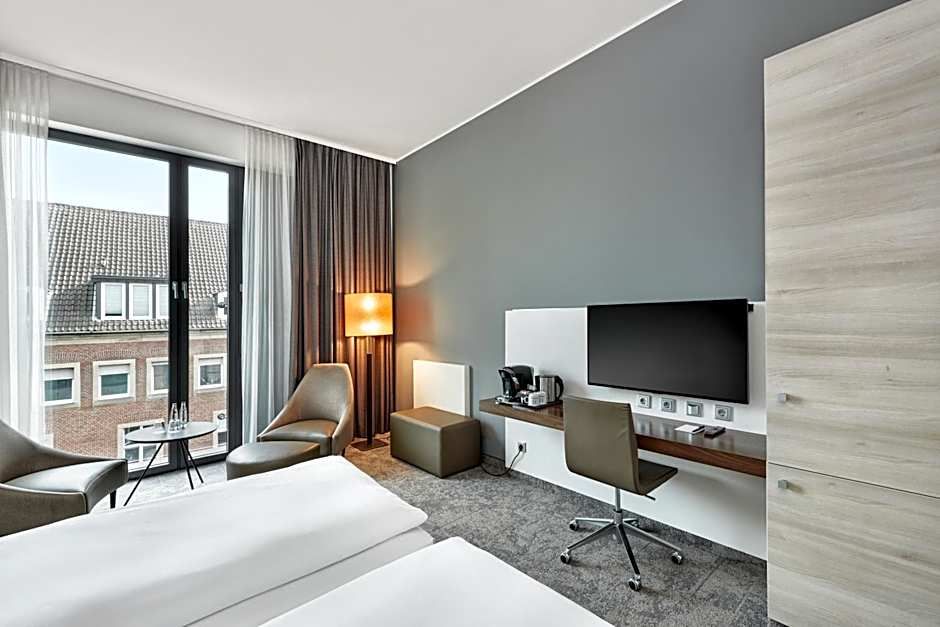 Helles Hotelzimmer mit zwei Einzelbetten, großer Fensterfront, Schreibtisch mit Stuhl und Fernseher, bequemen Sesseln und moderner, gemütlicher Ausstattung in neutralen Farben.