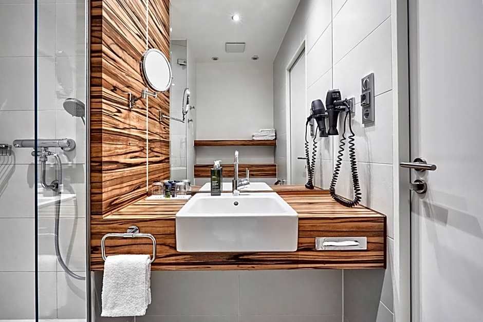 Modernes Hotelbadezimmer mit Holzakzenten, großem Spiegel, integriertem Waschbecken, Handtuchhalter, Haartrockner und einer Glasdusche. Sauber und funktional gestaltet für Komfort und praktische Nutzung.