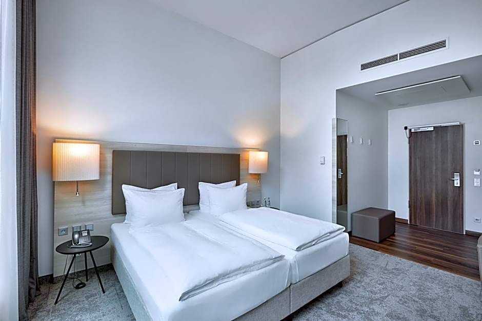 Modernes Hotelzimmer mit zwei Einzelbetten, beleuchteten Wandlampen, neutraler Farbgebung und Holzfußboden im Eingangsbereich, das eine ruhige und komfortable Atmosphäre für Geschäfts- und Freizeitreisende schafft.