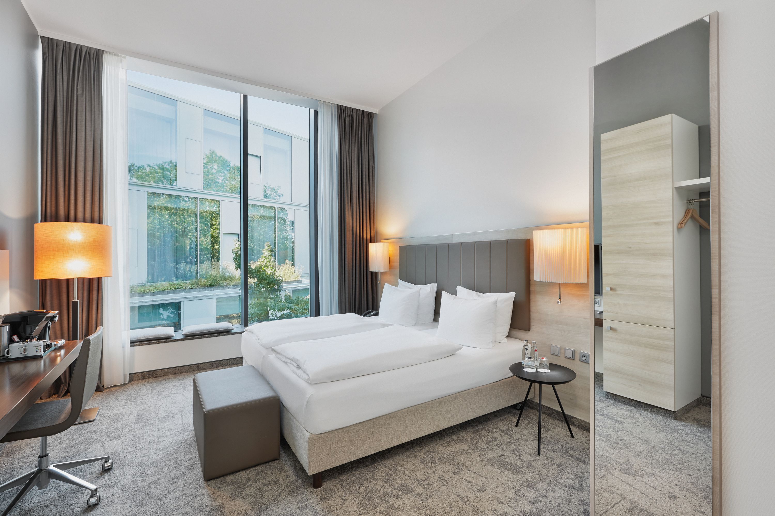 Modernes Doppelzimmer mit großem Fenster, Tageslicht, bequemen Betten, Arbeitsplatz und gemütlicher Beleuchtung für einen entspannten Aufenthalt.
