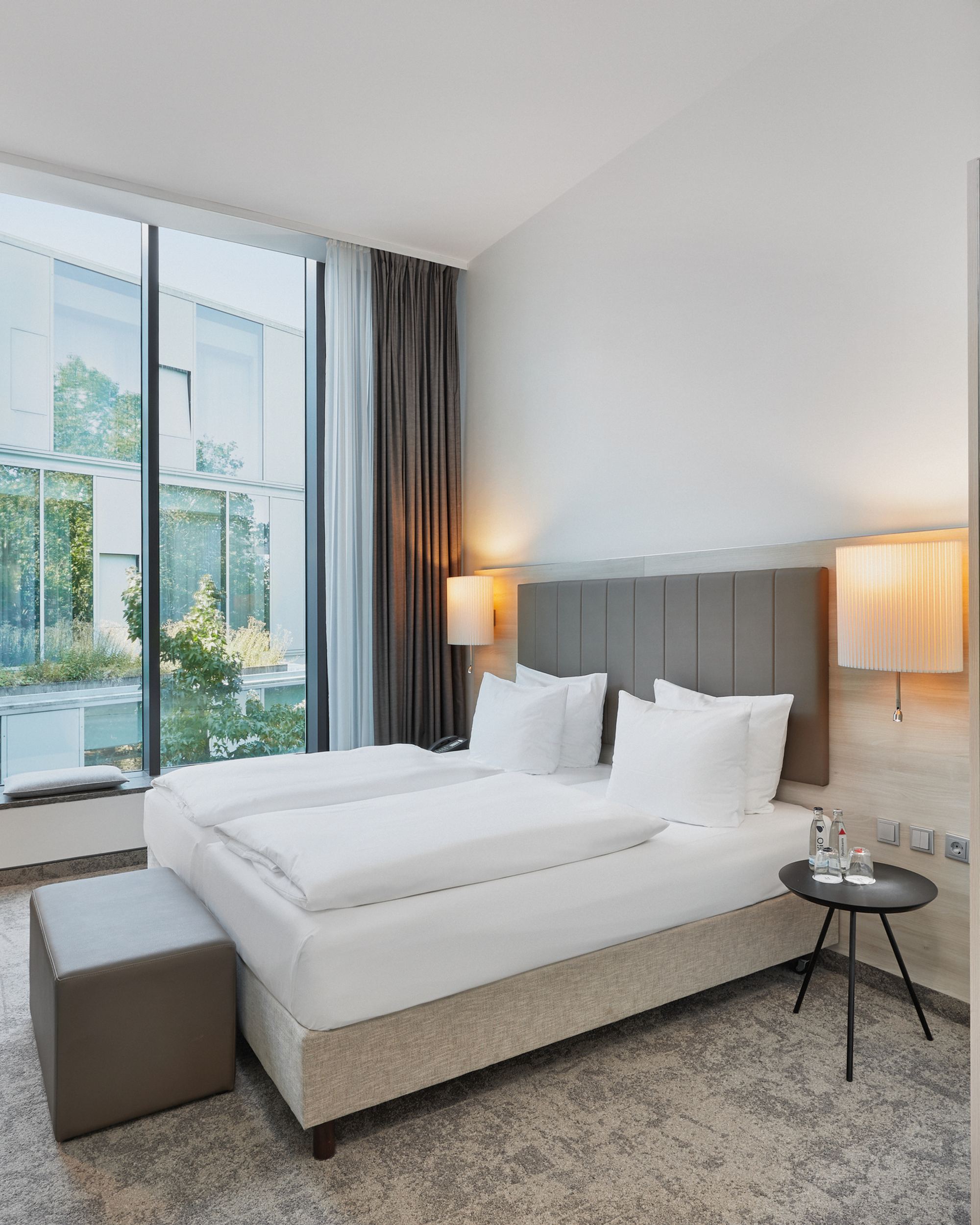 Modernes Doppelzimmer mit großem Fenster, Tageslicht, bequemen Betten, Arbeitsplatz und gemütlicher Beleuchtung für einen entspannten Aufenthalt.