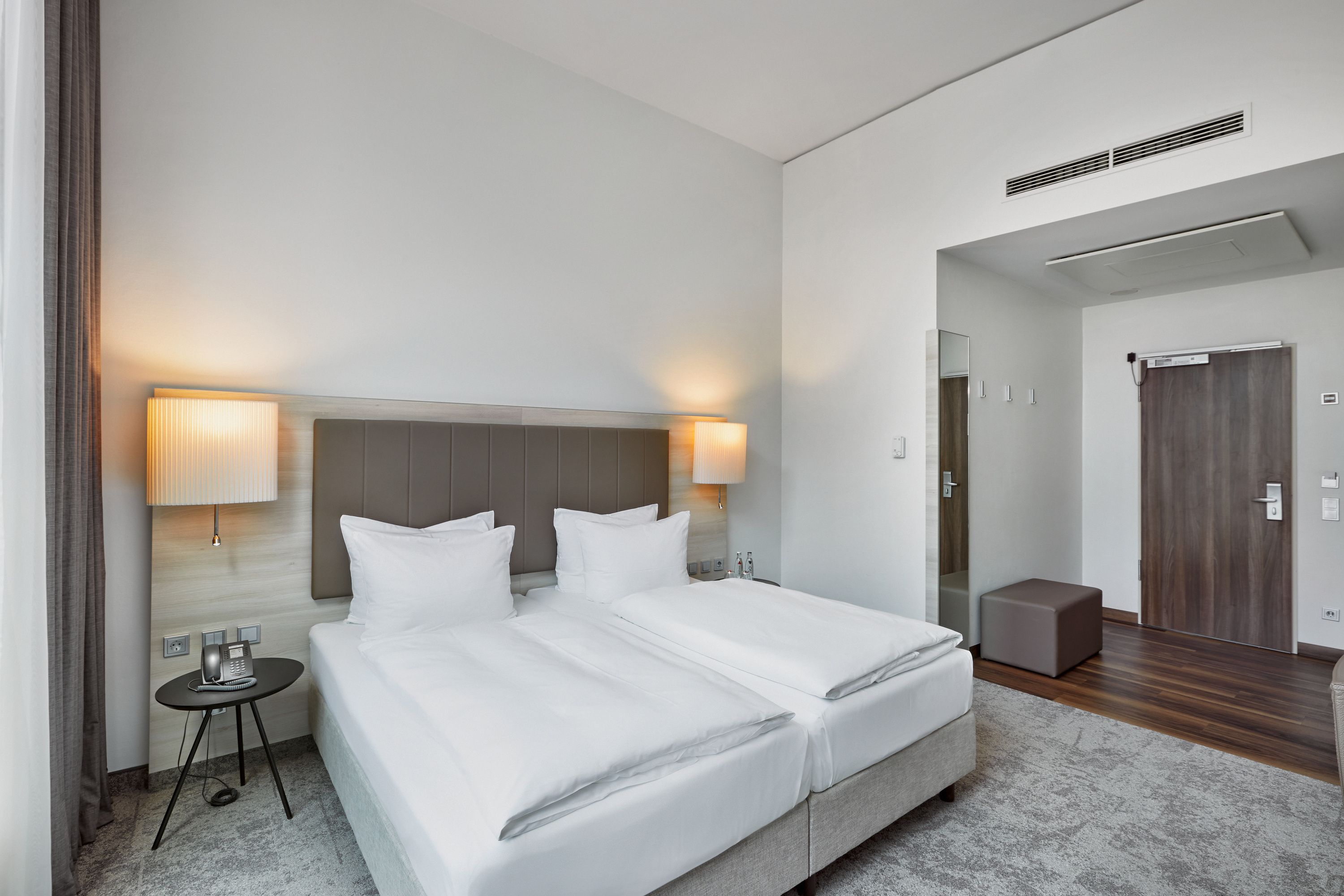 Modernes, helles Hotelzimmer mit zwei Einzelbetten, weicher Beleuchtung durch Wandlampen, schlichtem Design und ruhiger Atmosphäre, ideal für entspannte Übernachtung.