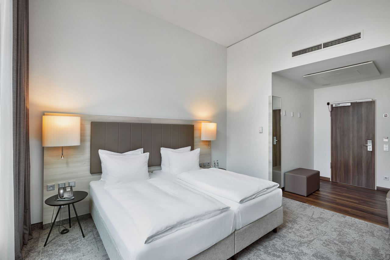 Modernes, helles Hotelzimmer mit zwei Einzelbetten, weicher Beleuchtung durch Wandlampen, schlichtem Design und ruhiger Atmosphäre, ideal für entspannte Übernachtung.