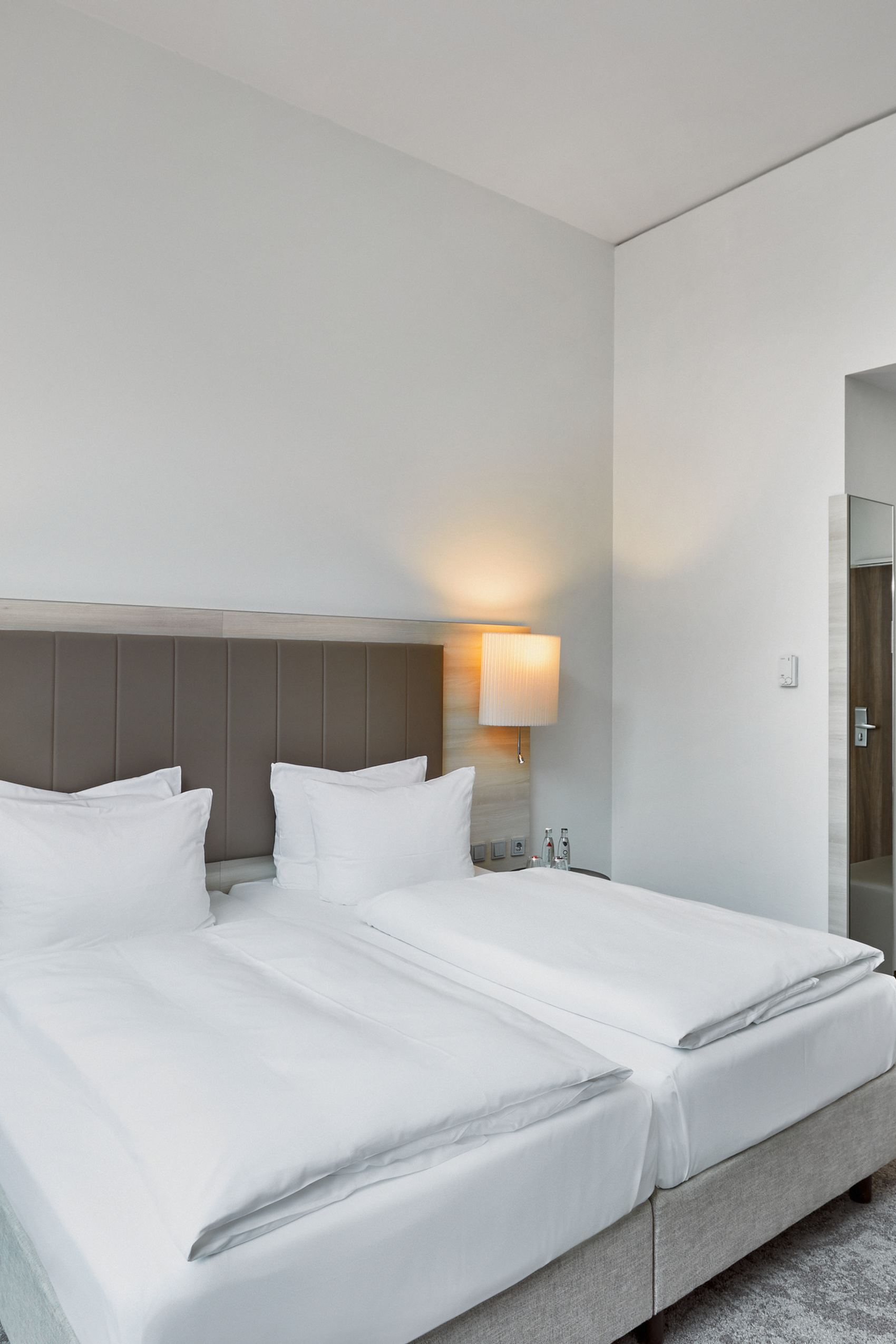 Modernes, helles Hotelzimmer mit zwei Einzelbetten, weicher Beleuchtung durch Wandlampen, schlichtem Design und ruhiger Atmosphäre, ideal für entspannte Übernachtung.