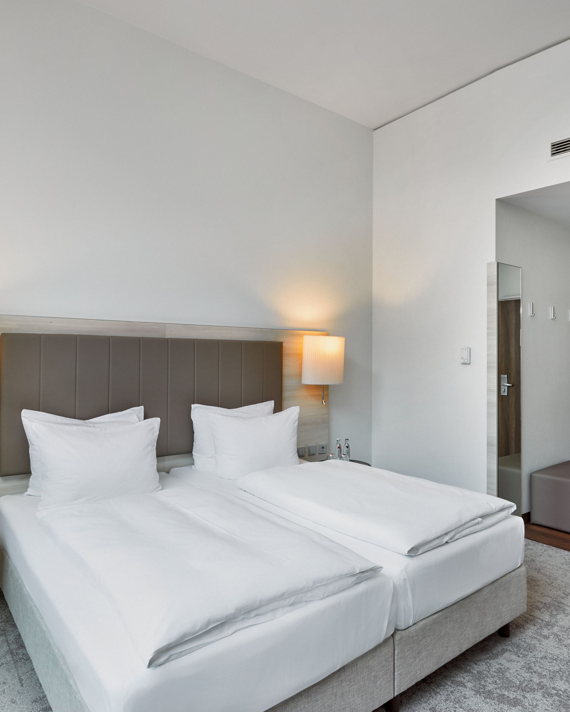 Modernes, helles Hotelzimmer mit zwei Einzelbetten, weicher Beleuchtung durch Wandlampen, schlichtem Design und ruhiger Atmosphäre, ideal für entspannte Übernachtung.