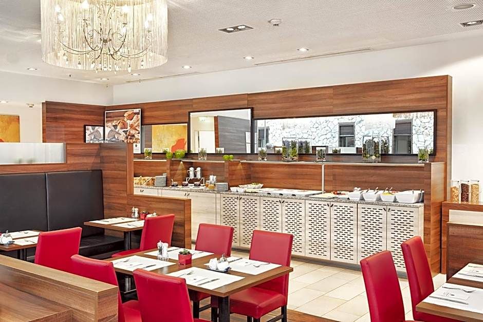 Helles, modernes Frühstücksrestaurant mit roten Stühlen, Holzakzenten und großem Buffetbereich, der verschiedene Speisen und Getränke anbietet. Gemütliche Atmosphäre für einen angenehmen Start in den Tag.