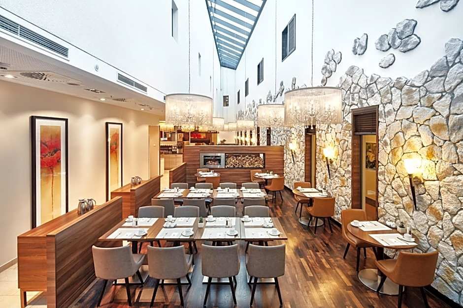 Helles, stilvolles Restaurant mit Holzboden, moderner Beleuchtung und Natursteinwänden. Mehrere gedeckte Tische und Stühle, große Fenster im Oberlicht sorgen für Tageslicht und ein einladendes Ambiente.