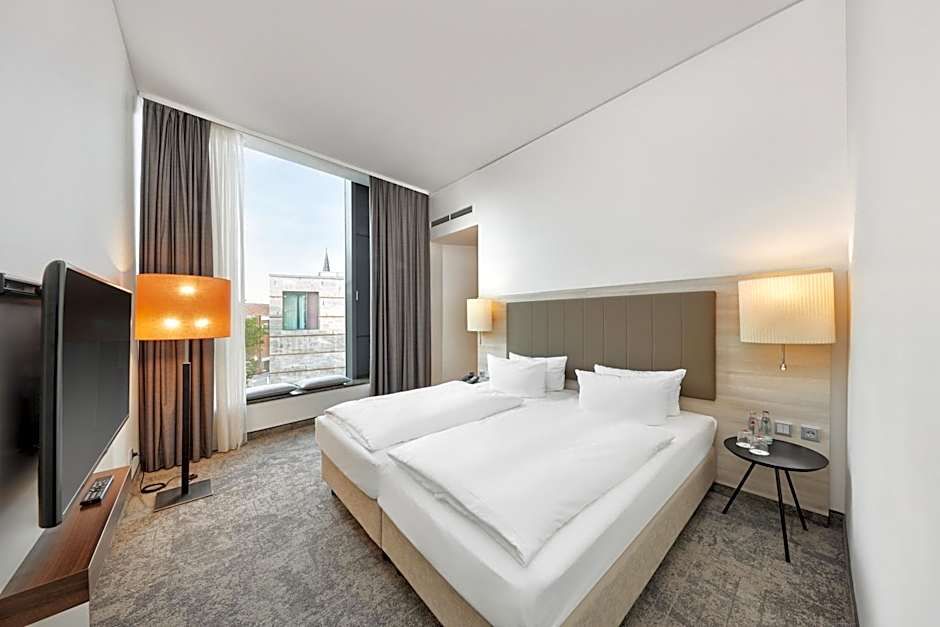 Helles, modernes Hotelzimmer mit zwei Einzelbetten, großen Fenstern mit Blick auf Stadtgebäude, neutralen Farben und gemütlicher Beleuchtung durch Steh- und Wandlampen für eine entspannte Atmosphäre.