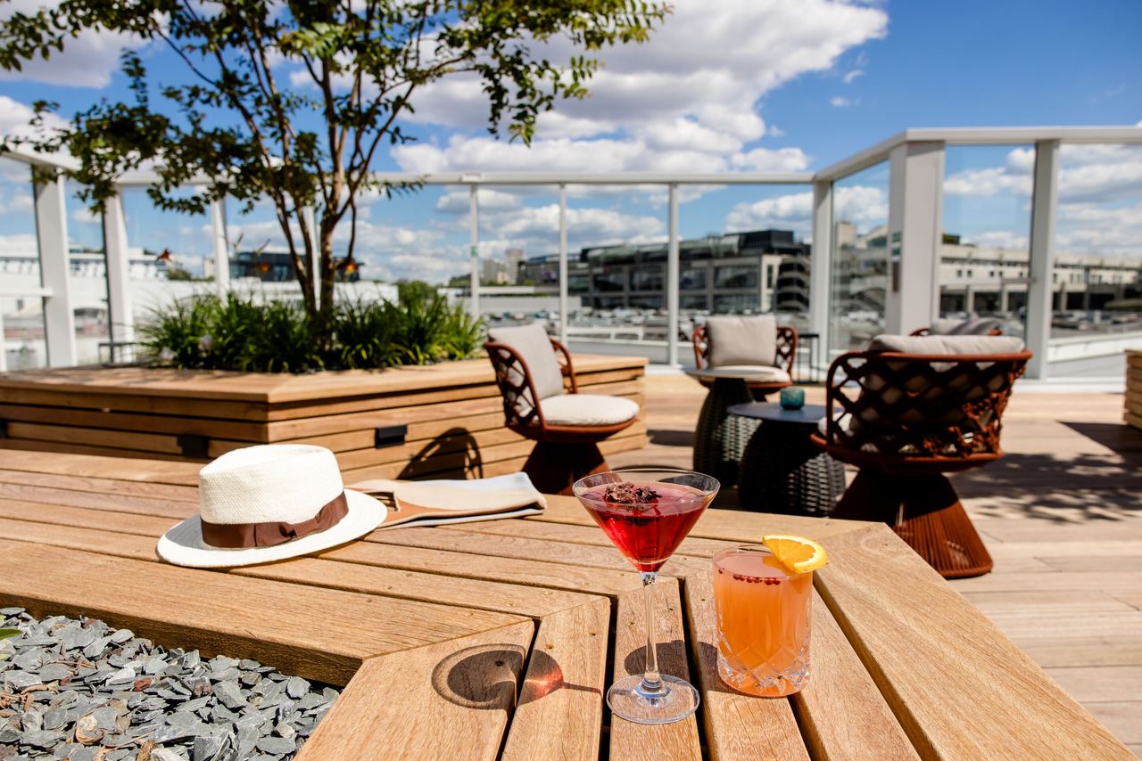Dachterrasse mit Holzdeck, bequemen Sitzmöbeln und Pflanzen, zwei farbenfrohe Cocktails auf dem Tisch, blauer Himmel mit weißen Wolken im Hintergrund