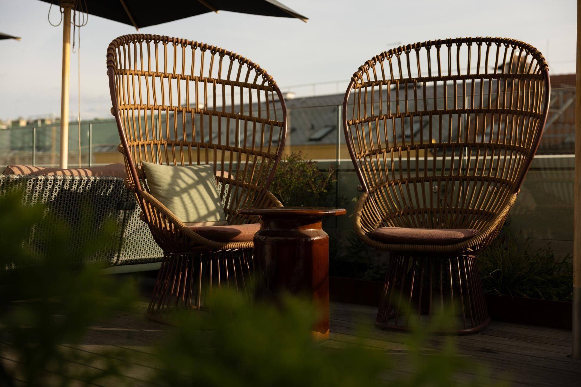 Zwei Rattanstühle mit gemütlichen Kissen stehen auf einer sonnigen Dachterrasse, umgeben von Pflanzen, ideal für entspannte Momente mit Ausblick.