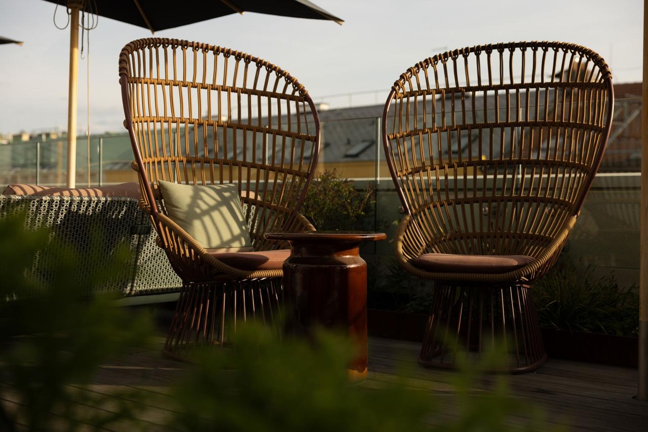 Zwei Rattanstühle mit gemütlichen Kissen stehen auf einer sonnigen Dachterrasse, umgeben von Pflanzen, ideal für entspannte Momente mit Ausblick.