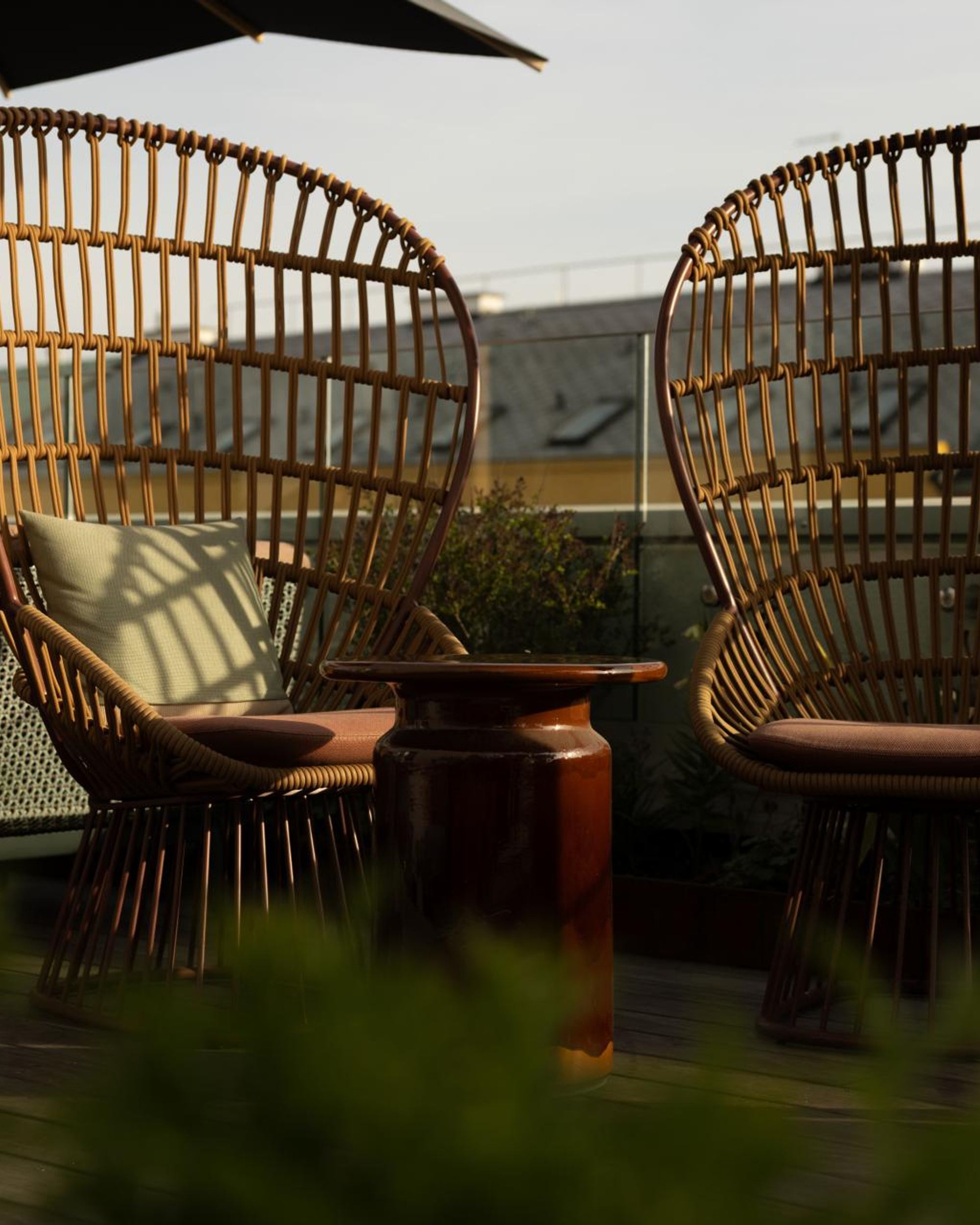 Zwei Rattanstühle mit gemütlichen Kissen stehen auf einer sonnigen Dachterrasse, umgeben von Pflanzen, ideal für entspannte Momente mit Ausblick.