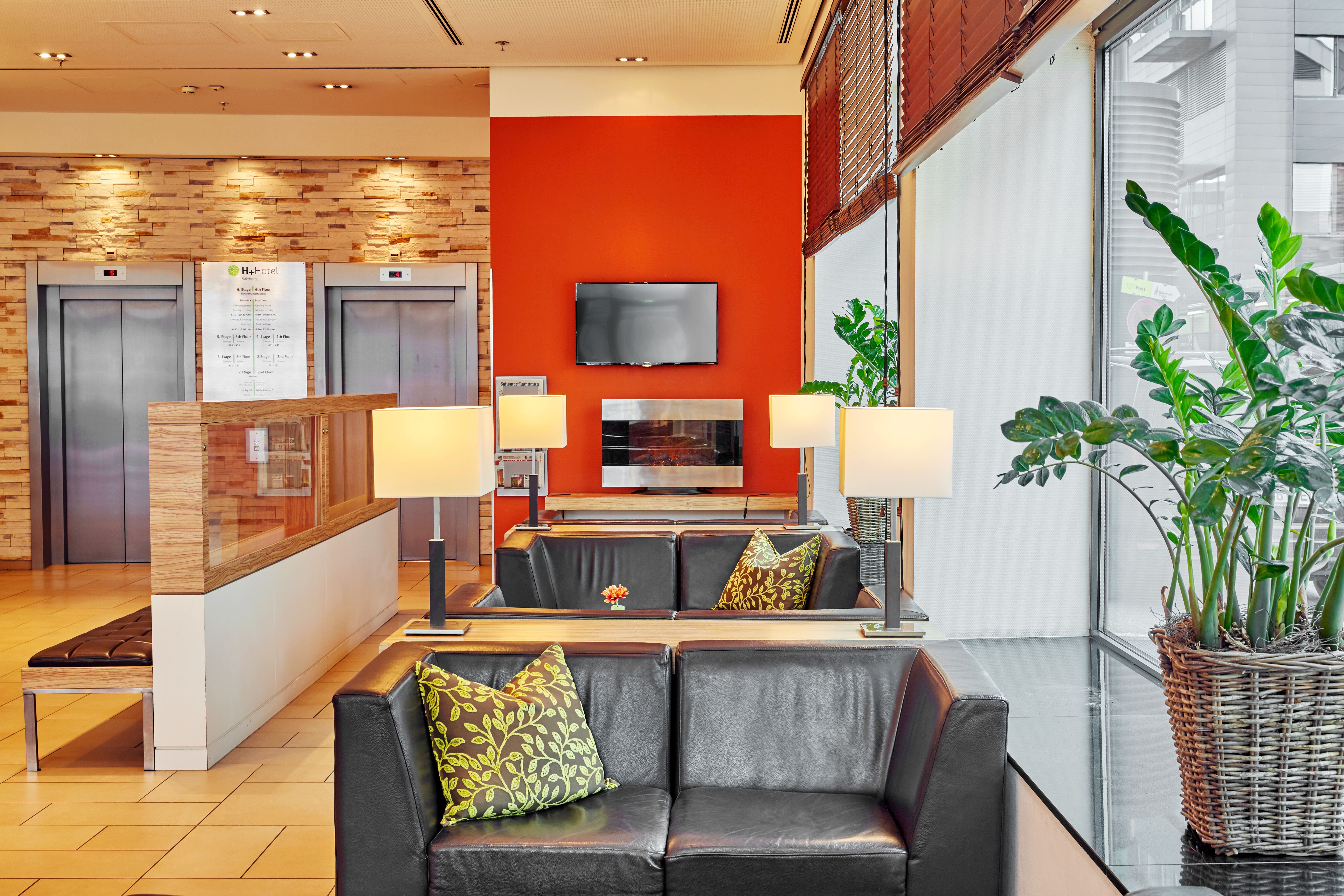 Moderne Hotellobby mit gemütlichem Sitzecke, Ledersofas und dekorativen Kissen. Große Fenster sorgen für viel Tageslicht, Pflanzen setzen grüne Akzente. Warme Beleuchtung und ein Bildschirm an der roten Akzentwand schaffen ein einladendes Ambiente. Aufzugsschacht mit zwei Aufzügen im Hintergrund.
