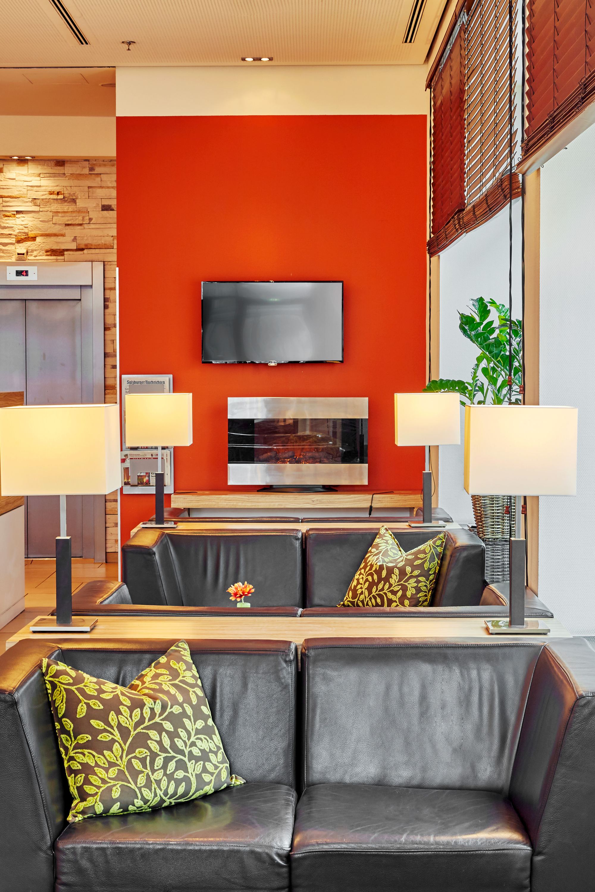 Moderne Hotellobby mit gemütlichem Sitzecke, Ledersofas und dekorativen Kissen. Große Fenster sorgen für viel Tageslicht, Pflanzen setzen grüne Akzente. Warme Beleuchtung und ein Bildschirm an der roten Akzentwand schaffen ein einladendes Ambiente. Aufzugsschacht mit zwei Aufzügen im Hintergrund.