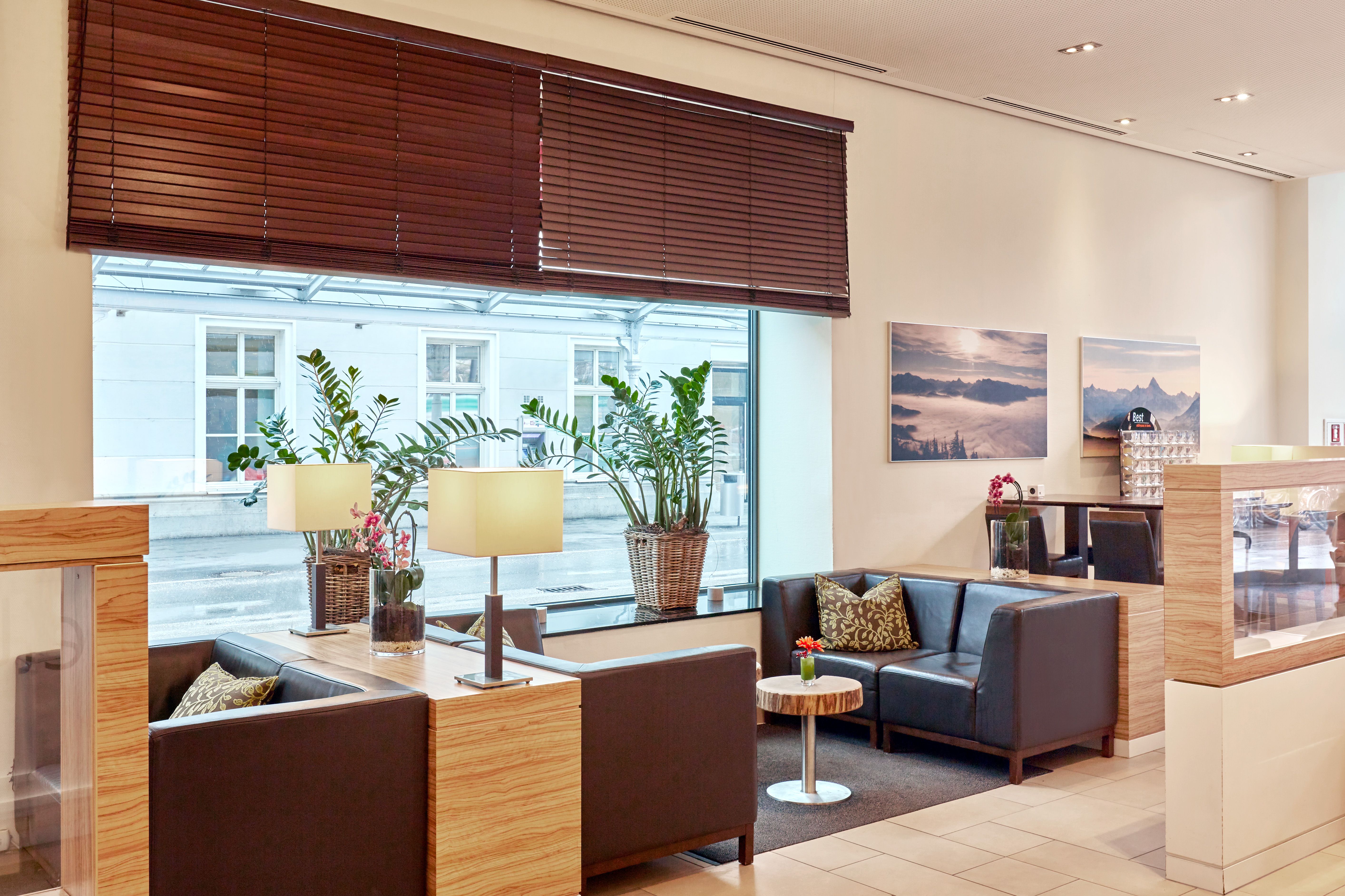Gemütliche Lounge mit modernen Ledersofas, kleinen Holztischen, grünen Pflanzen und sanfter Beleuchtung in warmem, einladendem Ambiente. Große Fenster bieten Tageslicht und urbane Aussicht.