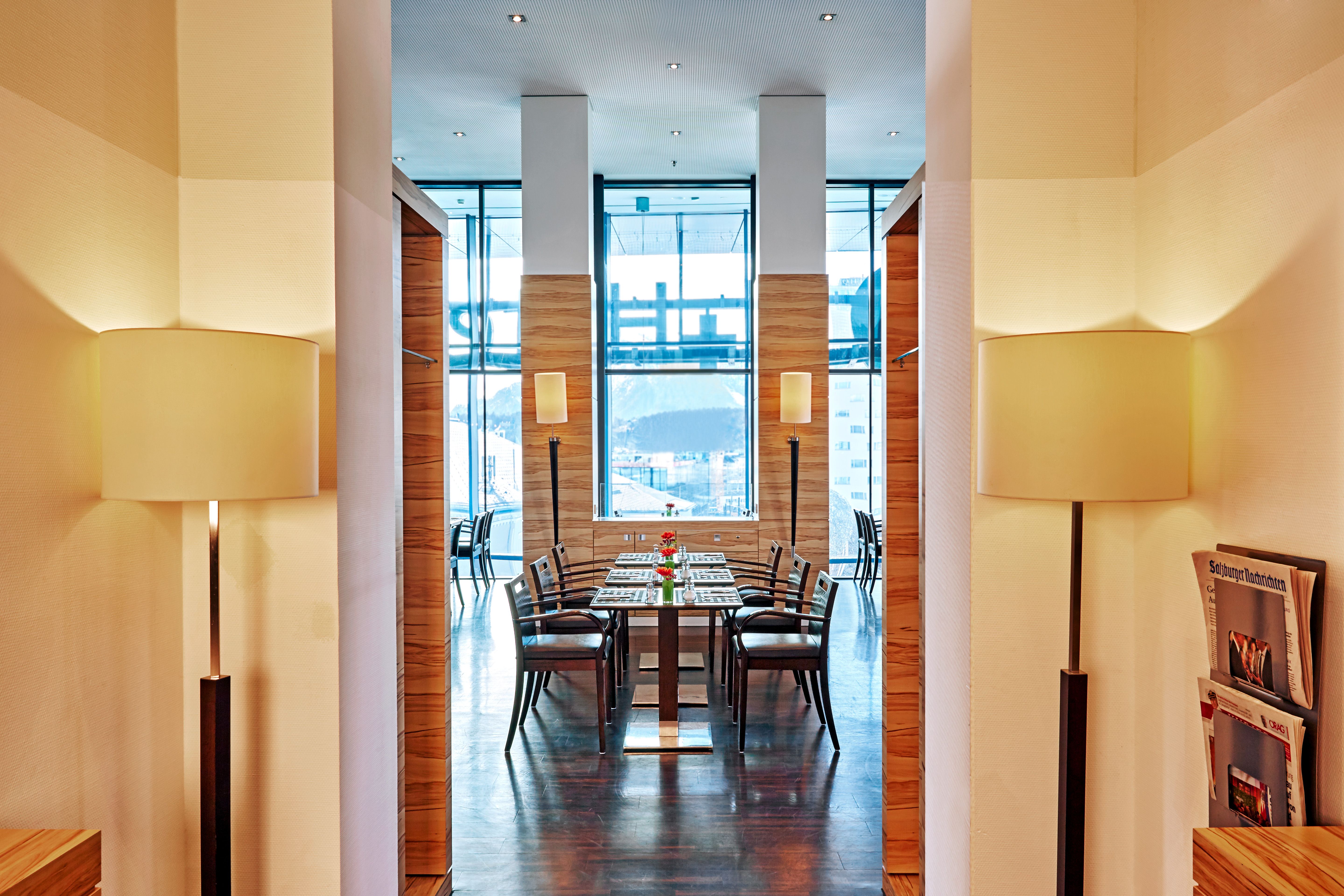 Elegantes Restaurant mit Holzvertäfelung, modernen Stühlen und gedecktem Tisch. Große Fenster bieten viel Tageslicht und Ausblick. Warme Beleuchtung durch Stehlampen schafft gemütliche Atmosphäre.