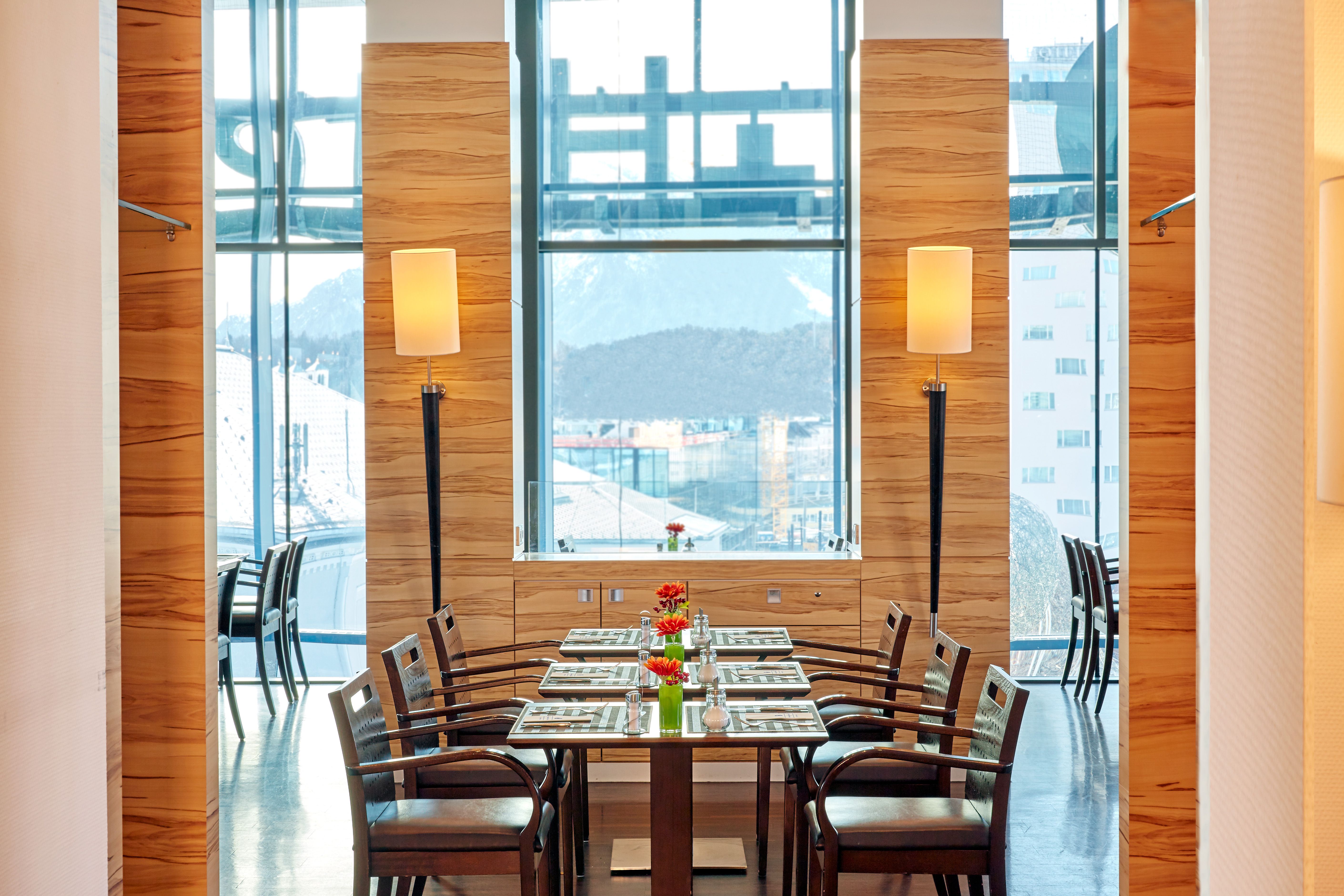 Modernes, helles Restaurant mit holzvertäfelten Wänden, großen Fenstern und Bergblick. Ein Tisch ist elegant mit Tellern, Gläsern und roten Blumen dekoriert, um gemütliches Speisen in stilvollem Ambiente zu ermöglichen.