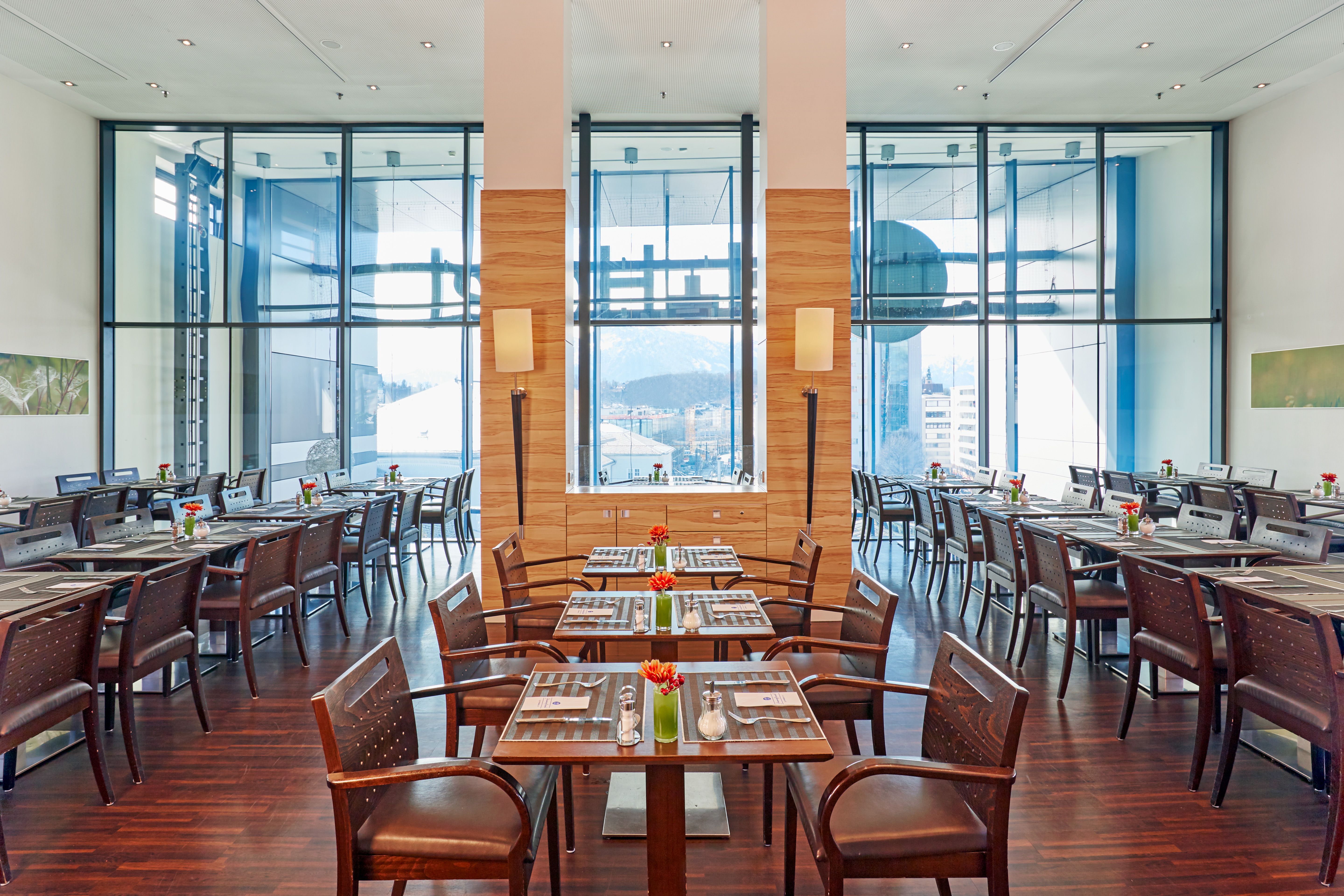 Helles, modernes Restaurant mit großen Fenstern, eleganten Holzmöbeln und gedeckten Tischen, die einladend für Gäste gestaltet sind. Blick auf die Stadt und Berge im Hintergrund. Angenehme Atmosphäre für entspannte Mahlzeiten.