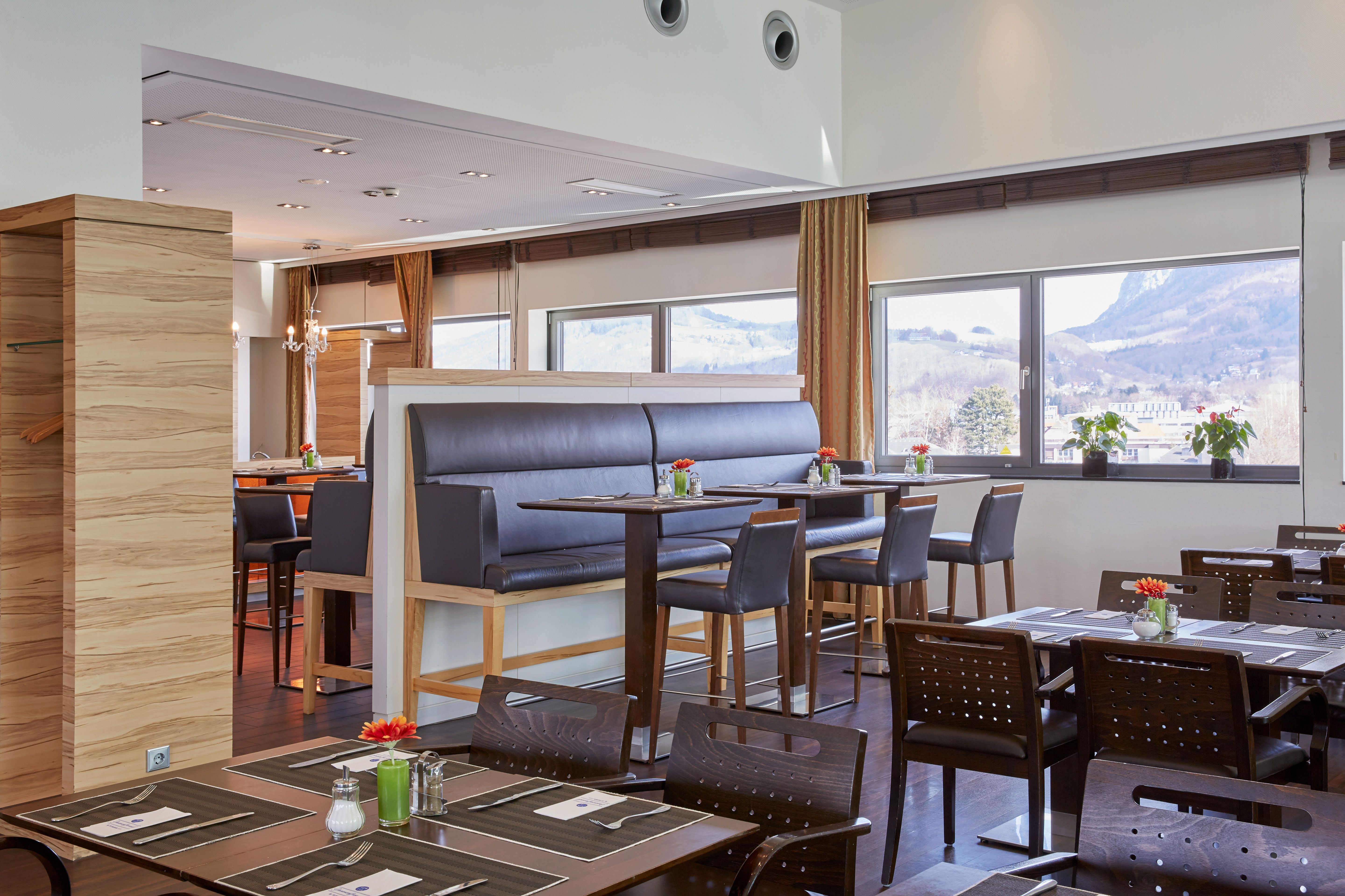 Helles Restaurant mit großen Fenstern, modernen Tischen und Stühlen, Sitzbänken und Blick auf gebirgige Landschaft, dekoriert mit kleinen Blumenvasen, einladend für entspannte Mahlzeiten.