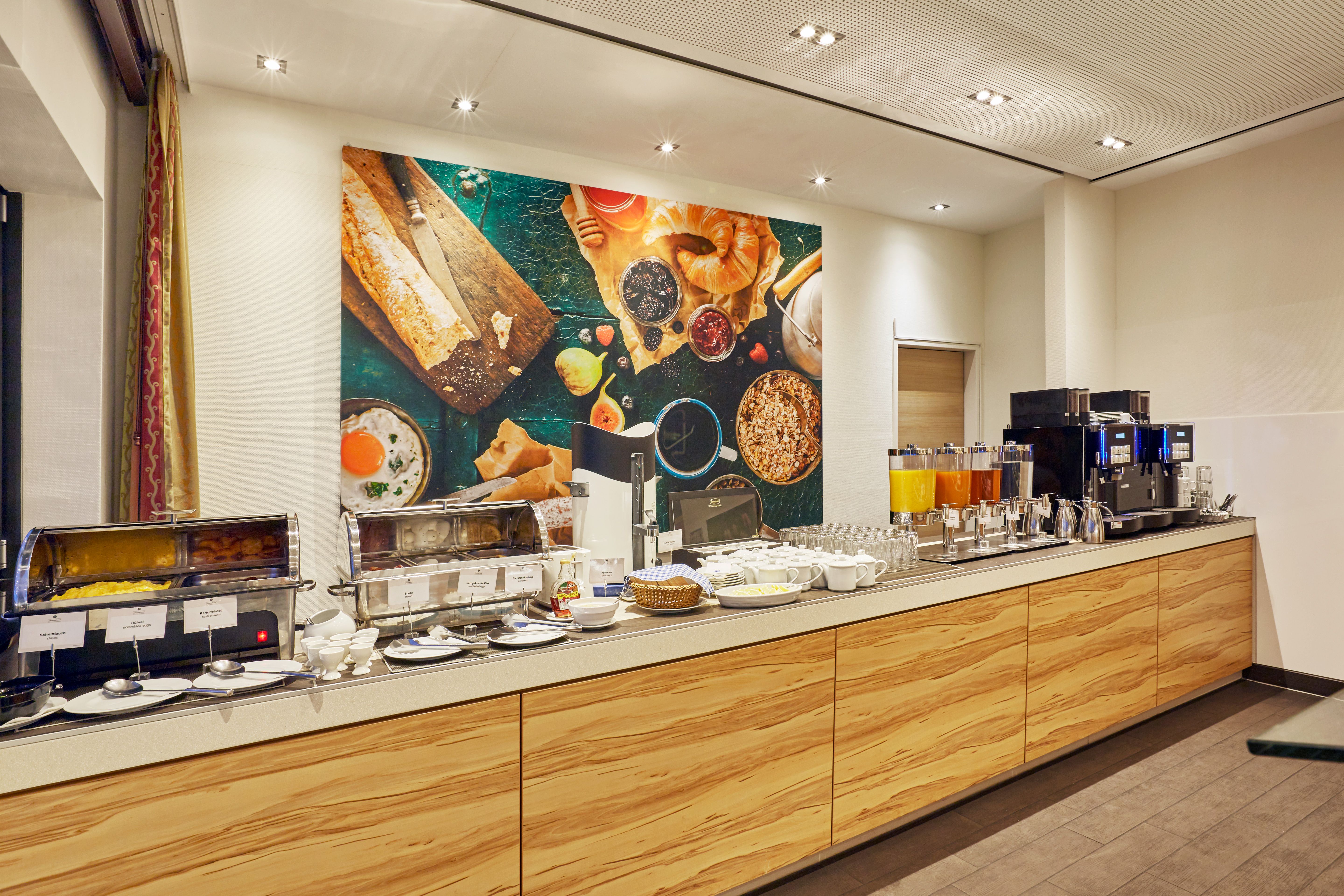 Buffetbereich mit vielfältigen Frühstücksangeboten, darunter Cerealien, Säfte, Kaffee und warme Speisen, in hellem, modernem Raum mit ansprechender Wanddekoration.