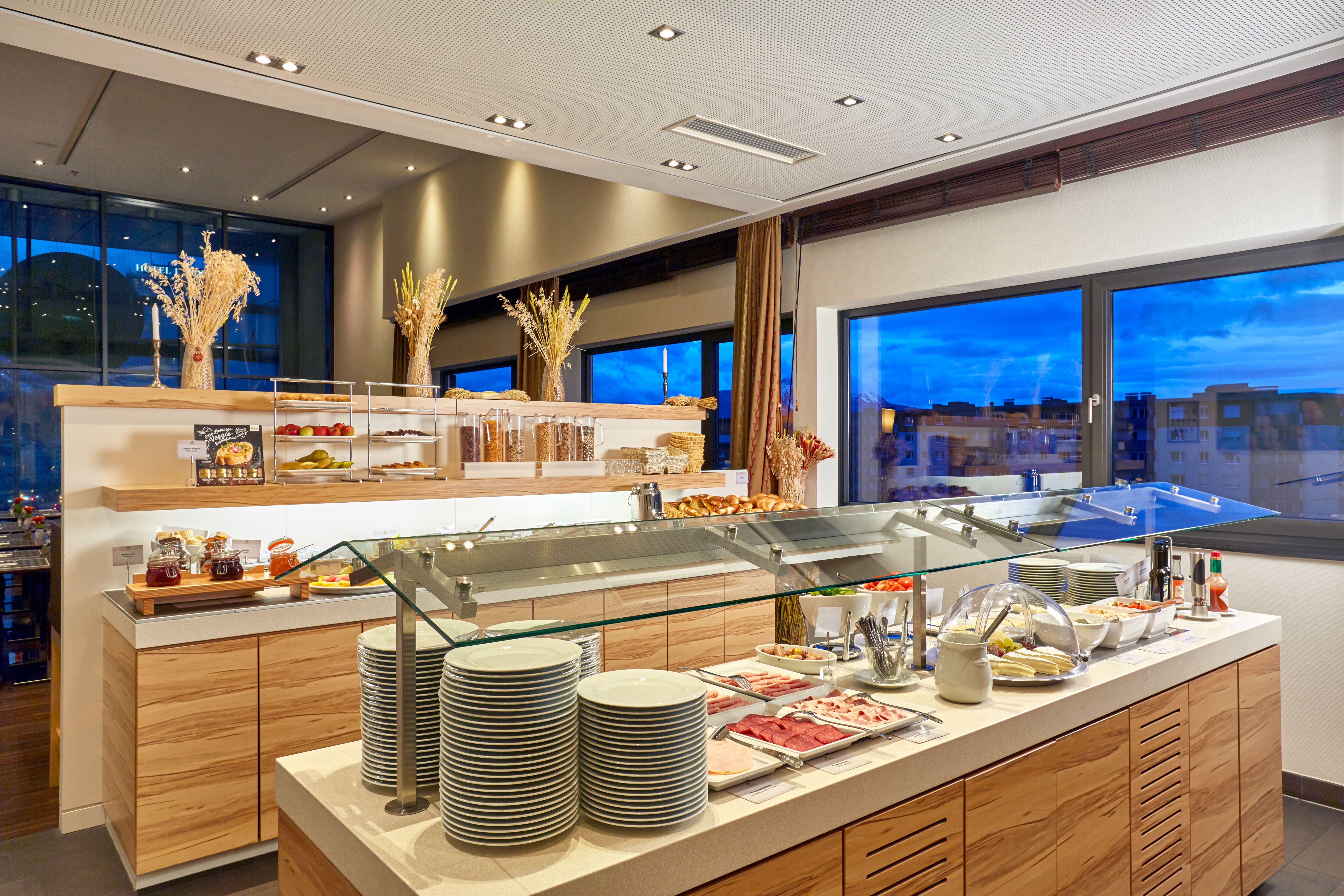 Buffetbereich mit vielfältigem Frühstücksangebot, darunter Wurst, Käse, Gebäck und Müsli, in modern eingerichteter Atmosphäre mit großen Fenstern und Blick auf die Stadt bei Abenddämmerung.