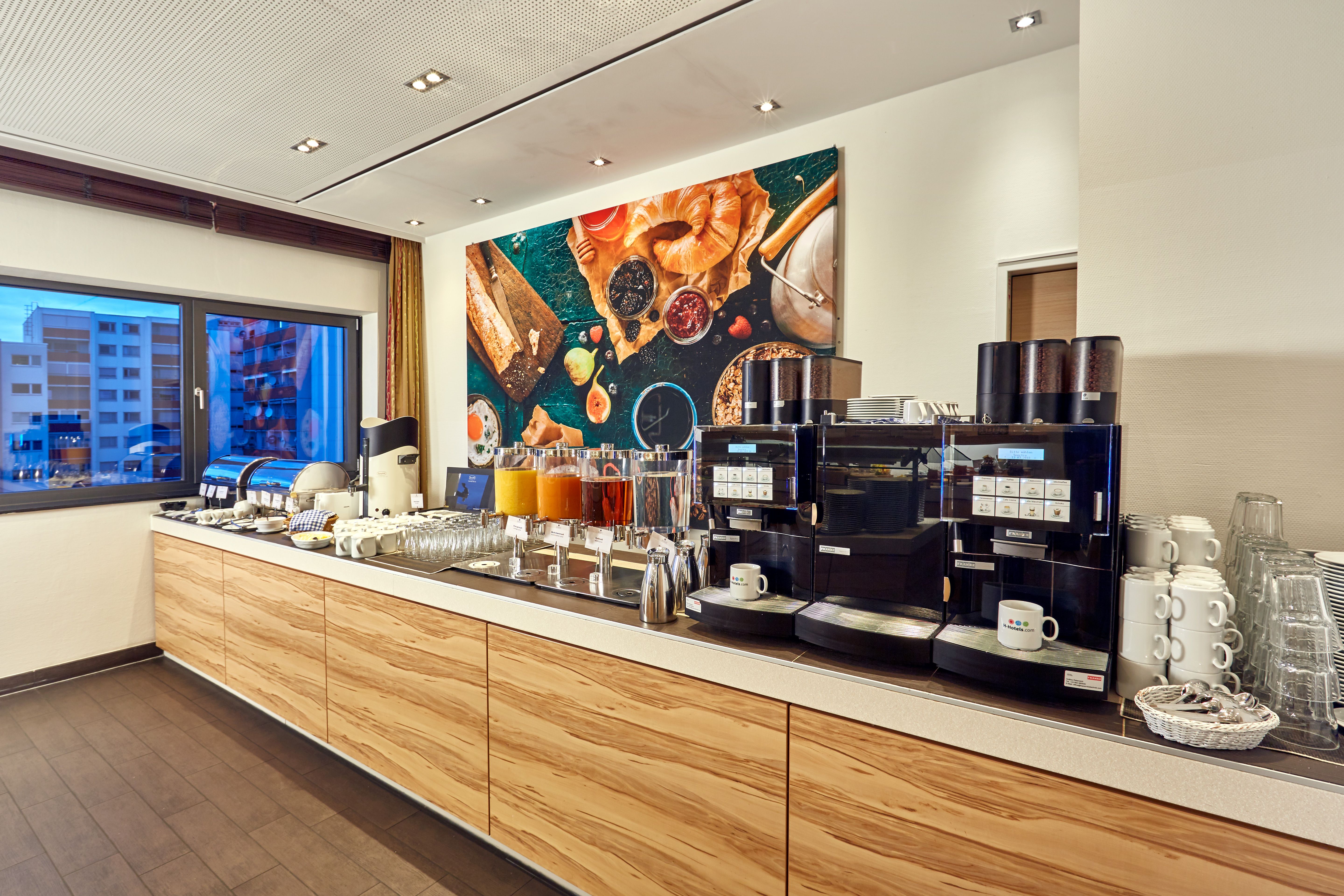 Moderne Frühstückstheke mit Kaffeevollautomaten, Teesortiment, Säften und leckeren Frühstückszutaten, helles Ambiente mit großem Fenster und ansprechender Wanddekoration aus Brot und Früchten.