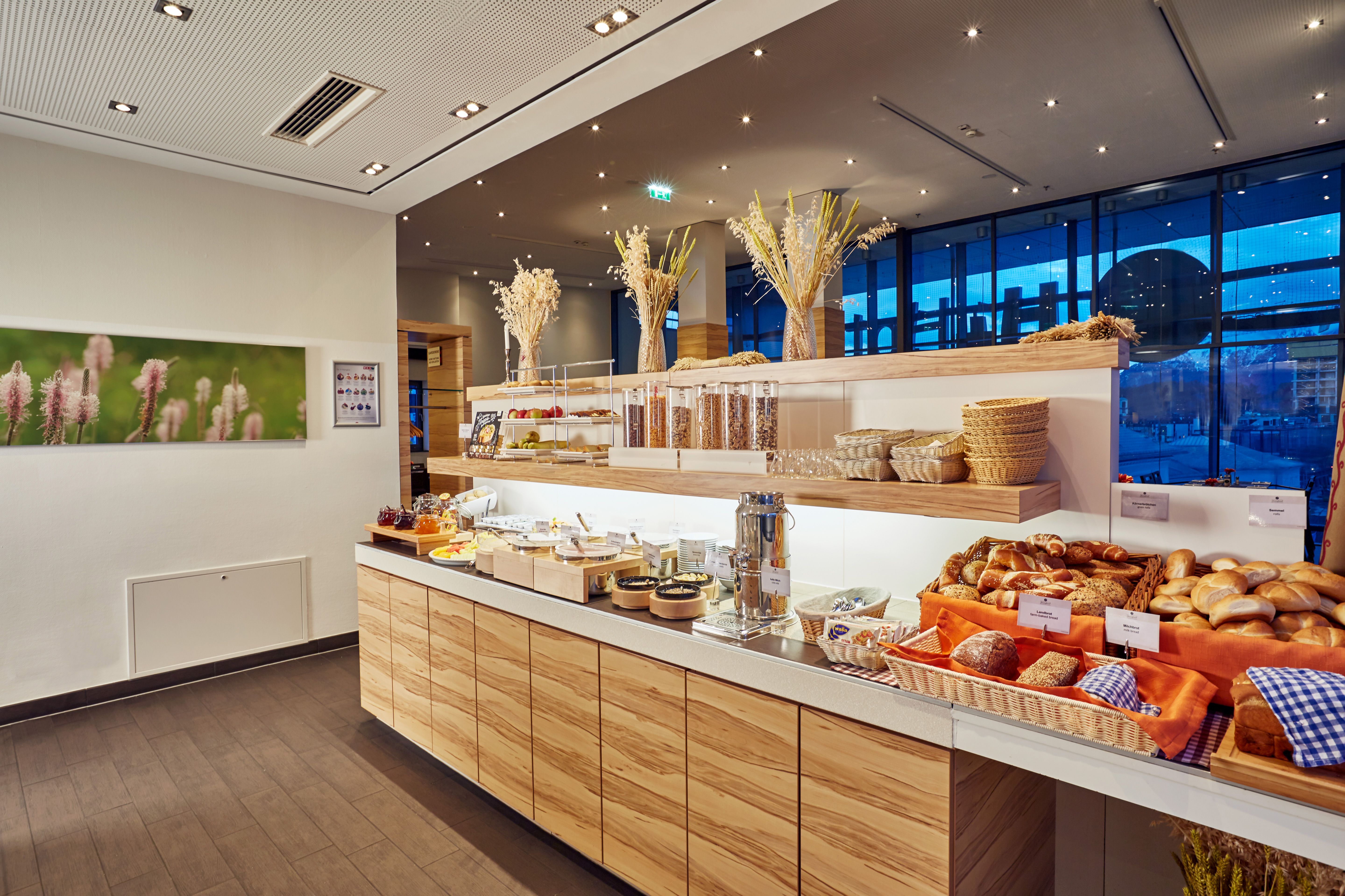 Frisches Frühstücksbuffet mit Brötchen, Croissants, Körnerbroten, verschiedenen Aufstrichen und Cerealien, liebevoll dekoriert in modernem, hellen Raum mit großem Fenster und Abenddämmerung draußen, gemütliche Atmosphäre für einen energiegeladenen Start in den Tag.