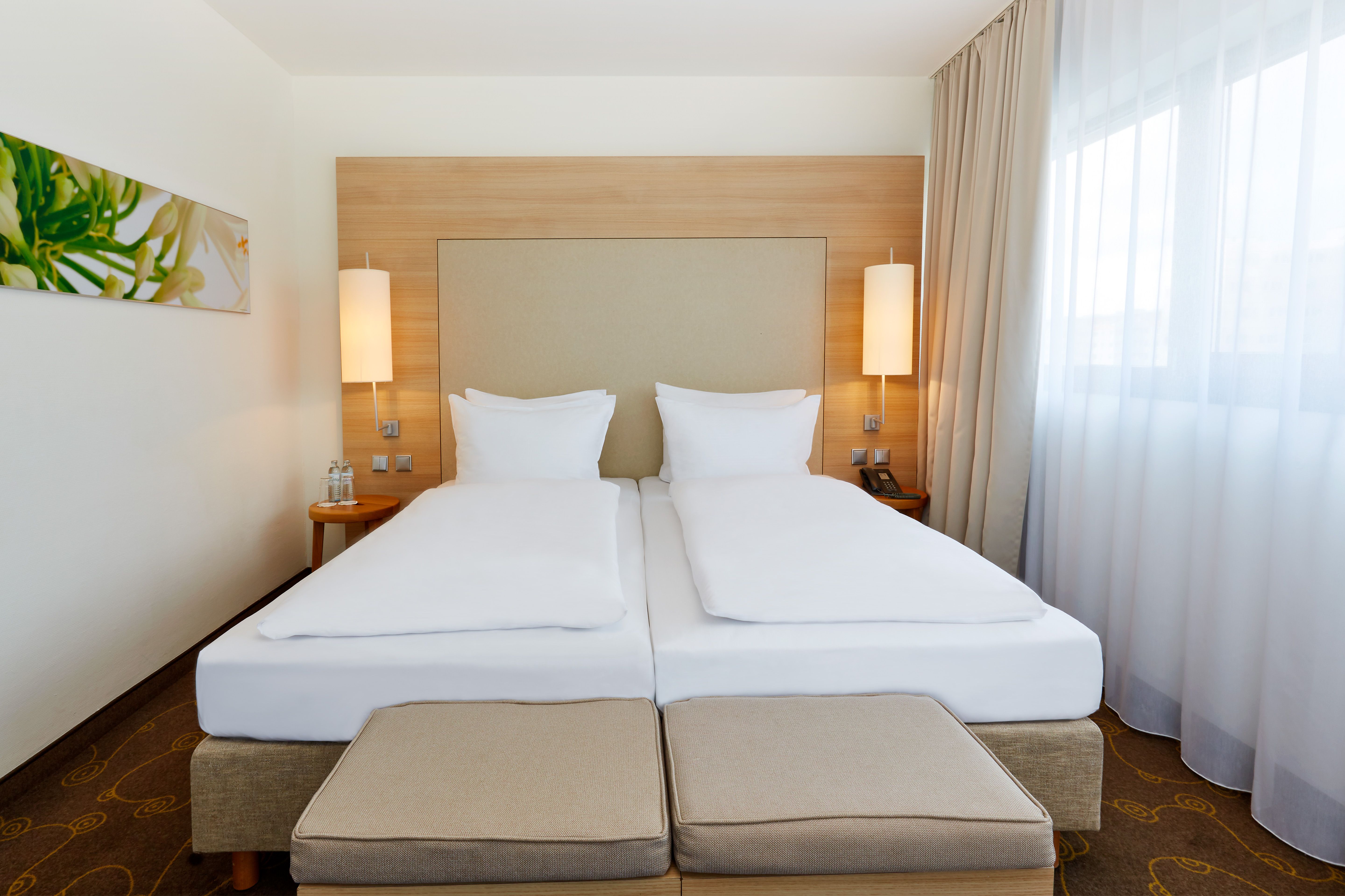 Modern eingerichtetes Hotelzimmer mit zwei Einzelbetten, hellen Bettwäsche, warmem Holzpaneel hinter dem Kopfteil, stilvollen Nachttischlampen und großen Fenstern mit Tageslicht.