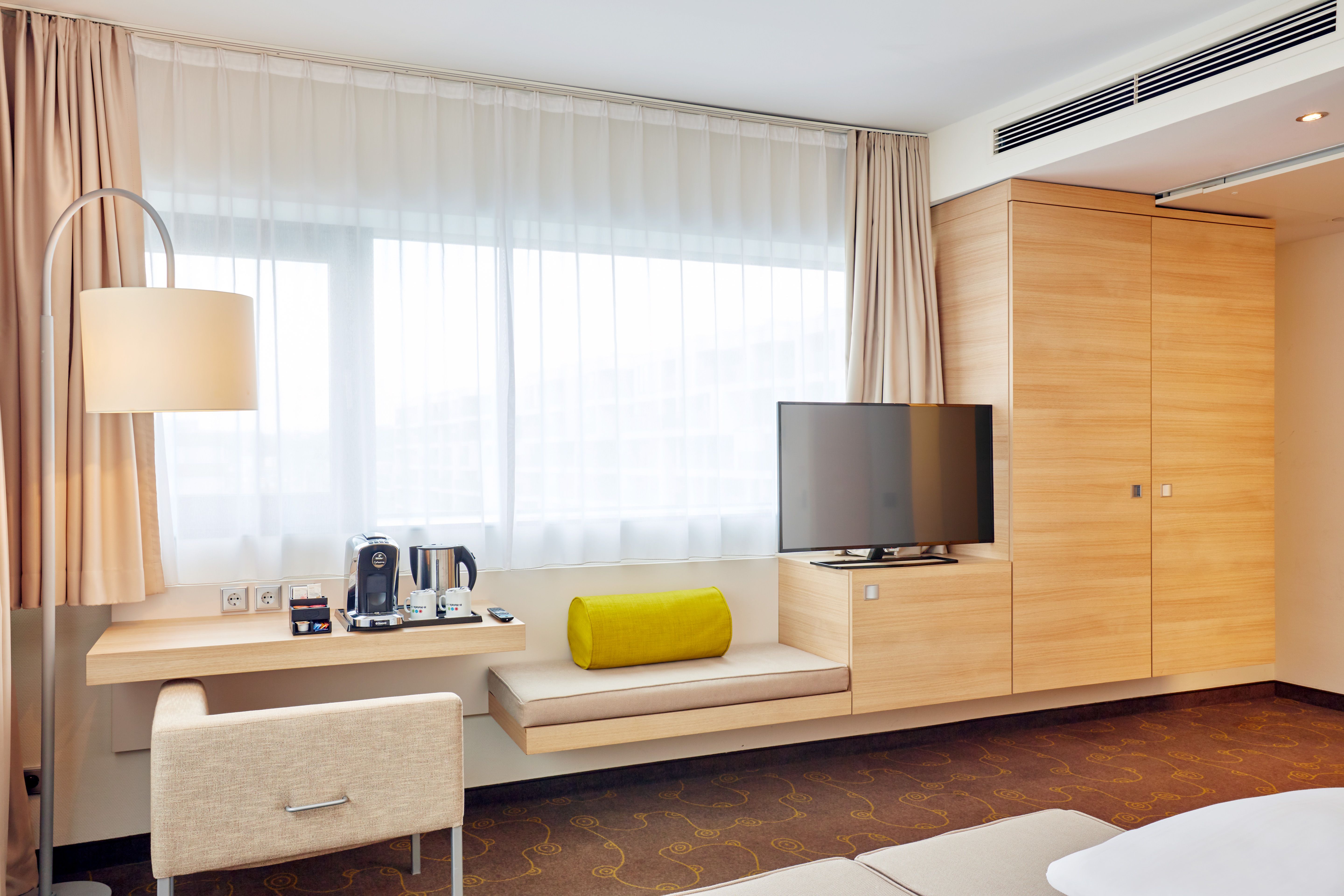 Helles Hotelzimmer mit großem Fenster, gemütlicher Sitzbank, Schreibtisch mit Kaffeemaschine, Flachbildfernseher und moderner Holzmöblierung für einen entspannten Aufenthalt.