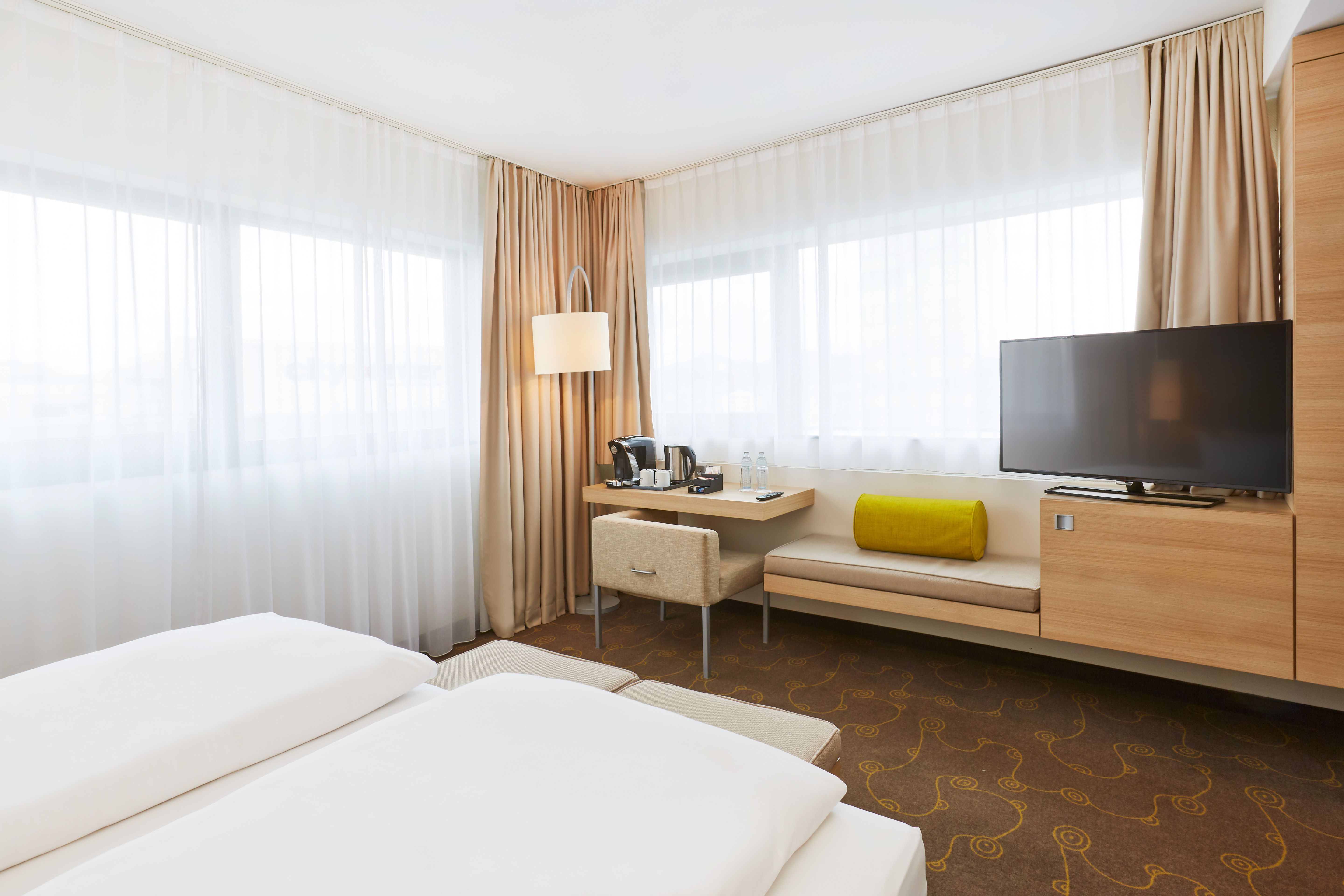 Helles, modernes Hotelzimmer mit zwei Betten, großem Fenster, Schreibtisch mit Kaffeemaschine, gemütlicher Sitzbank und Flachbildfernseher.