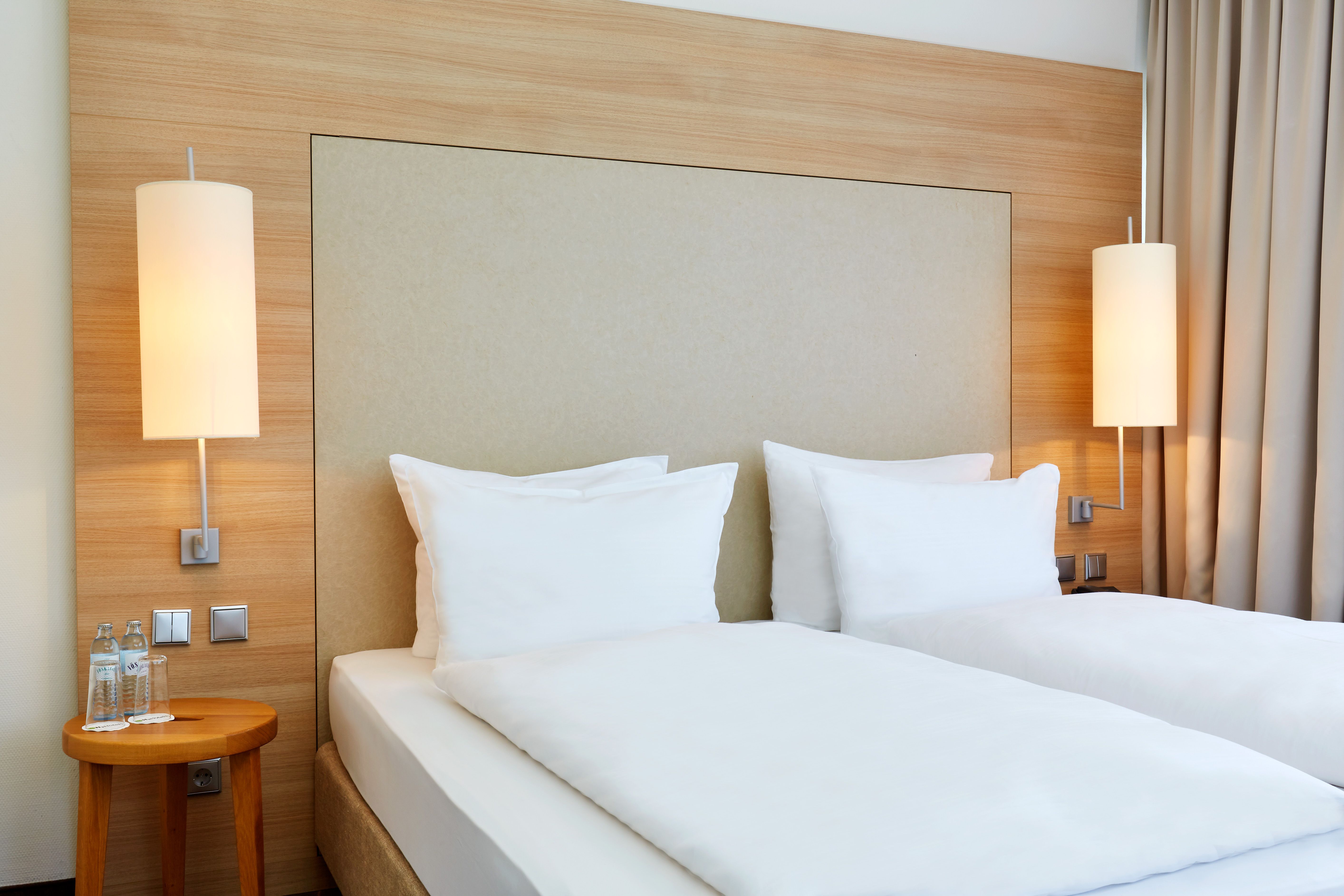 Zwei Einzelbetten mit weißen Bettdecken und Kissen in einem hellen, modernen Hotelzimmer, flankiert von zwei Wandlampen und einem kleinen Beistelltisch mit Wasserflaschen.