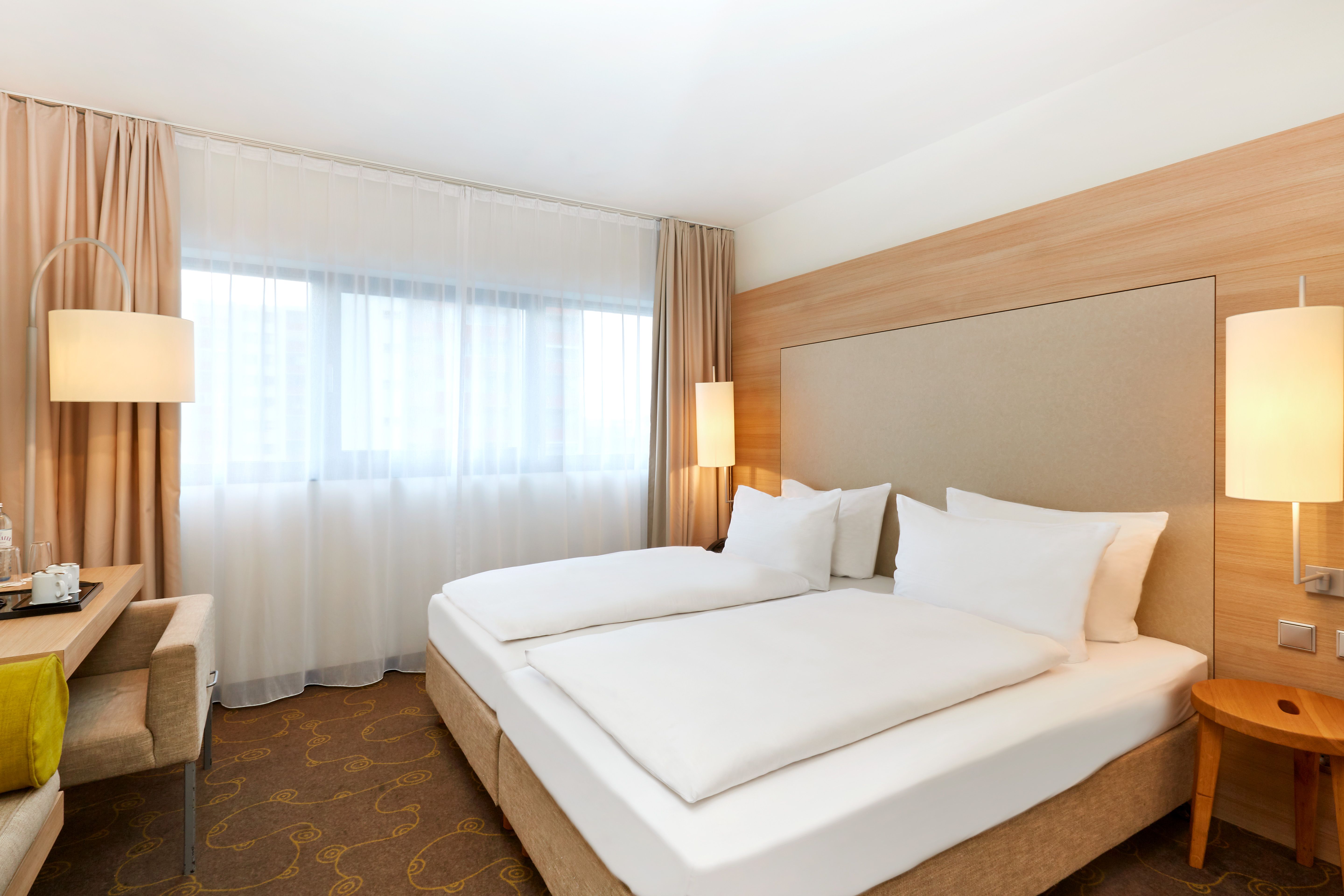 Modern eingerichtetes Doppelzimmer mit zwei Einzelbetten, warmem Licht, großen Fenstern und Schreibtisch, das eine ruhige und komfortable Atmosphäre für erholsamen Aufenthalt bietet.