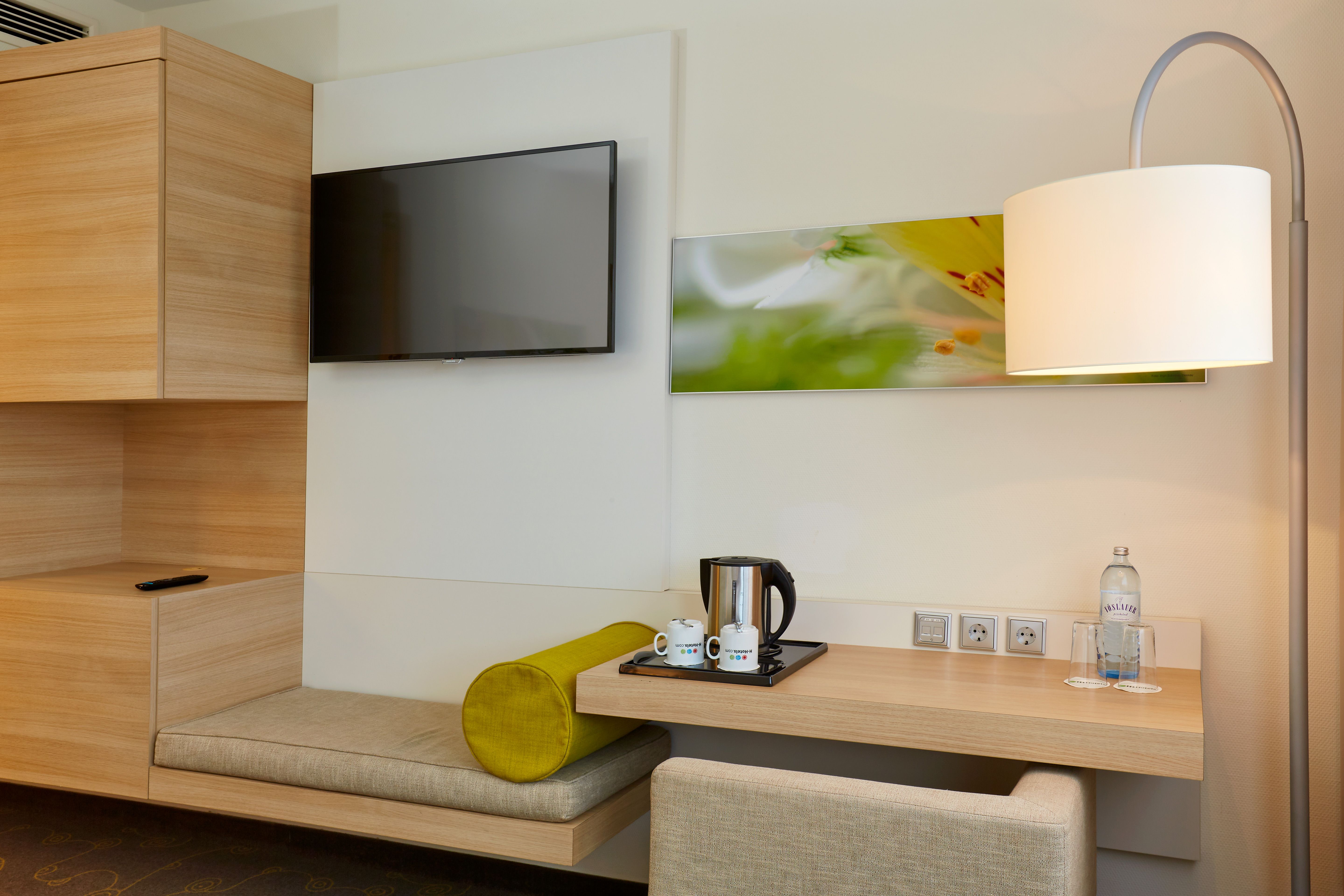 Modernes Hotelzimmer mit hellem Holzmobiliar, Sitzbank mit grünem Kissen, wandmontiertem Flachbildschirm, Schreibtisch mit Wasserflasche, Kaffeemaschine und Stehlampe für angenehmes Ambiente.