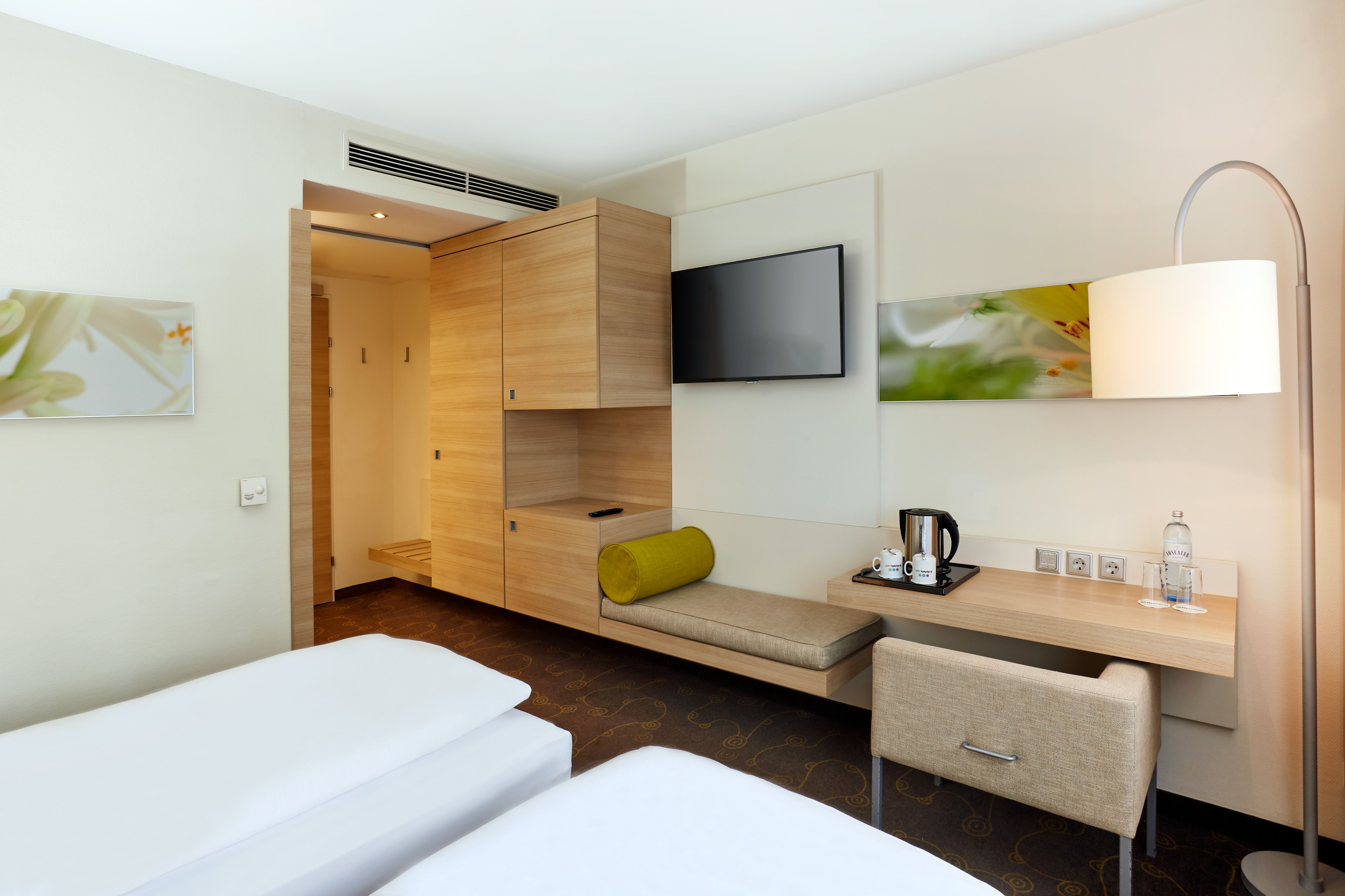 Modern eingerichtetes Hotelzimmer mit zwei getrennten Betten, hellem Holzmobiliar, Wand-TV, Sitzbank mit grünem Kissen und Teekocher. Helle, ruhige Atmosphäre lädt zum Erholen ein.