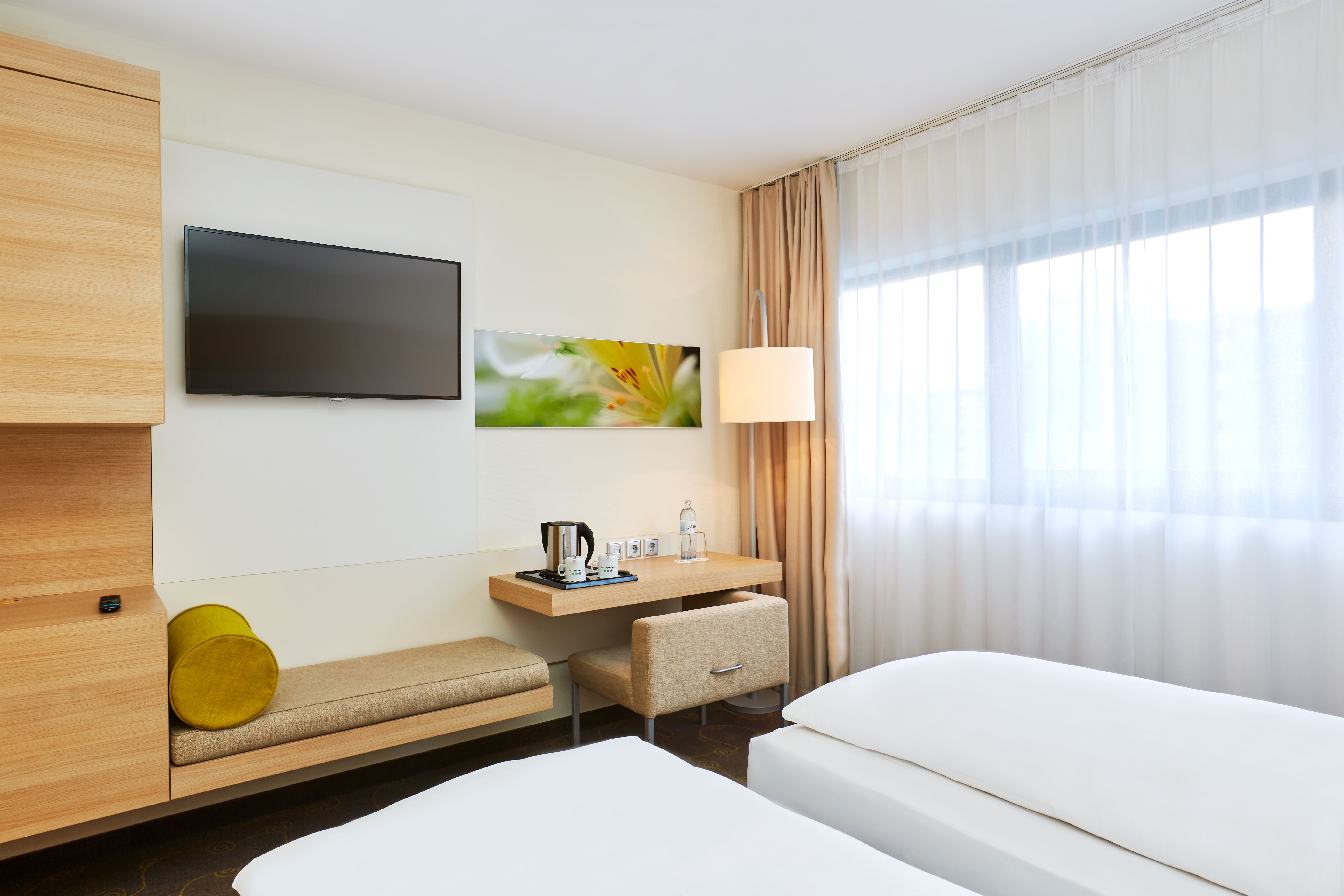 Hell eingerichtetes Hotelzimmer mit zwei Einzelbetten, großem Fenster mit Vorhängen, Sitzbank am Schreibtisch und moderner Wanddekoration für eine ruhige, komfortable Atmosphäre.