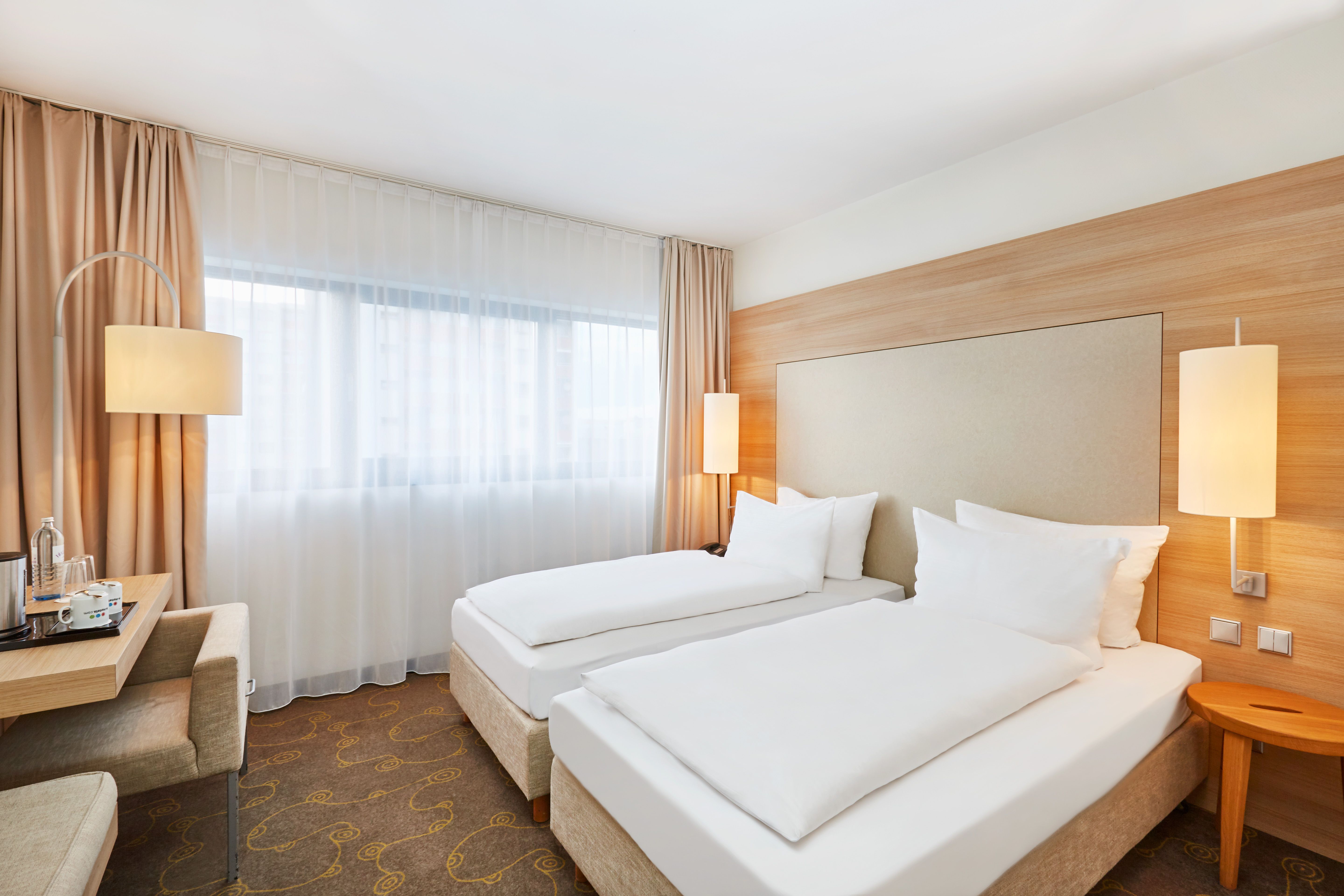 Helles Hotelzimmer mit zwei getrennten Betten, warmem Holzdesign, großen Fenstern mit Vorhängen und stimmungsvoller Beleuchtung für eine ruhige und komfortable Übernachtung.