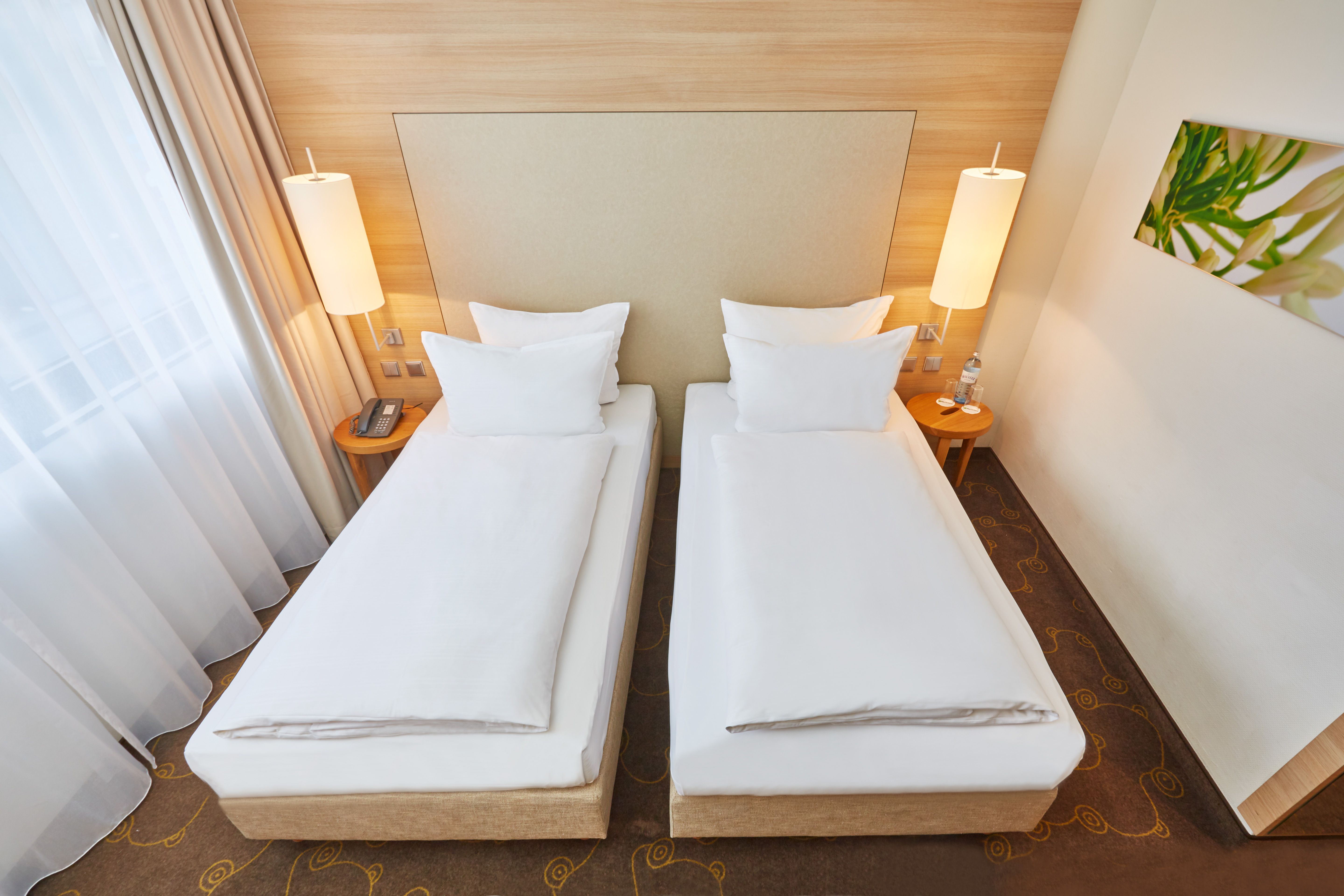 Zwei Einzelbetten mit weißen Bettlaken in einem hellen, modernen Hotelzimmer, seitlich je eine Nachttischlampe und schlichtes Wandbild, Tageslicht durch Gardinenfenster.