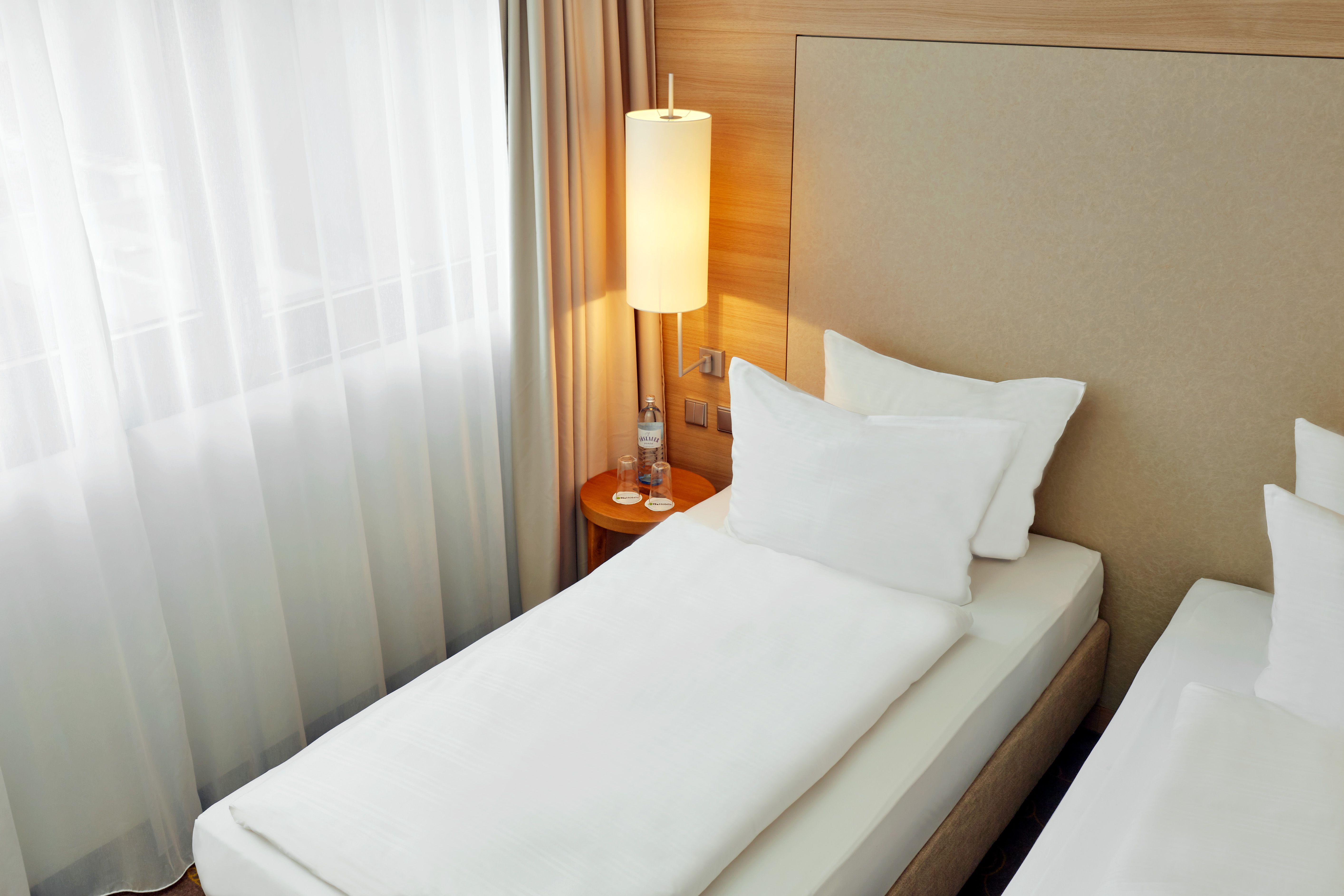 Helles Hotelzimmer mit zwei Einzelbetten, weißen Bettwäsche, einem Nachttisch mit Wasserflasche und modernem hängenden Lampenschirm, natürliche Lichtquelle durch ein großes Fenster mit halbtransparenten Vorhängen.