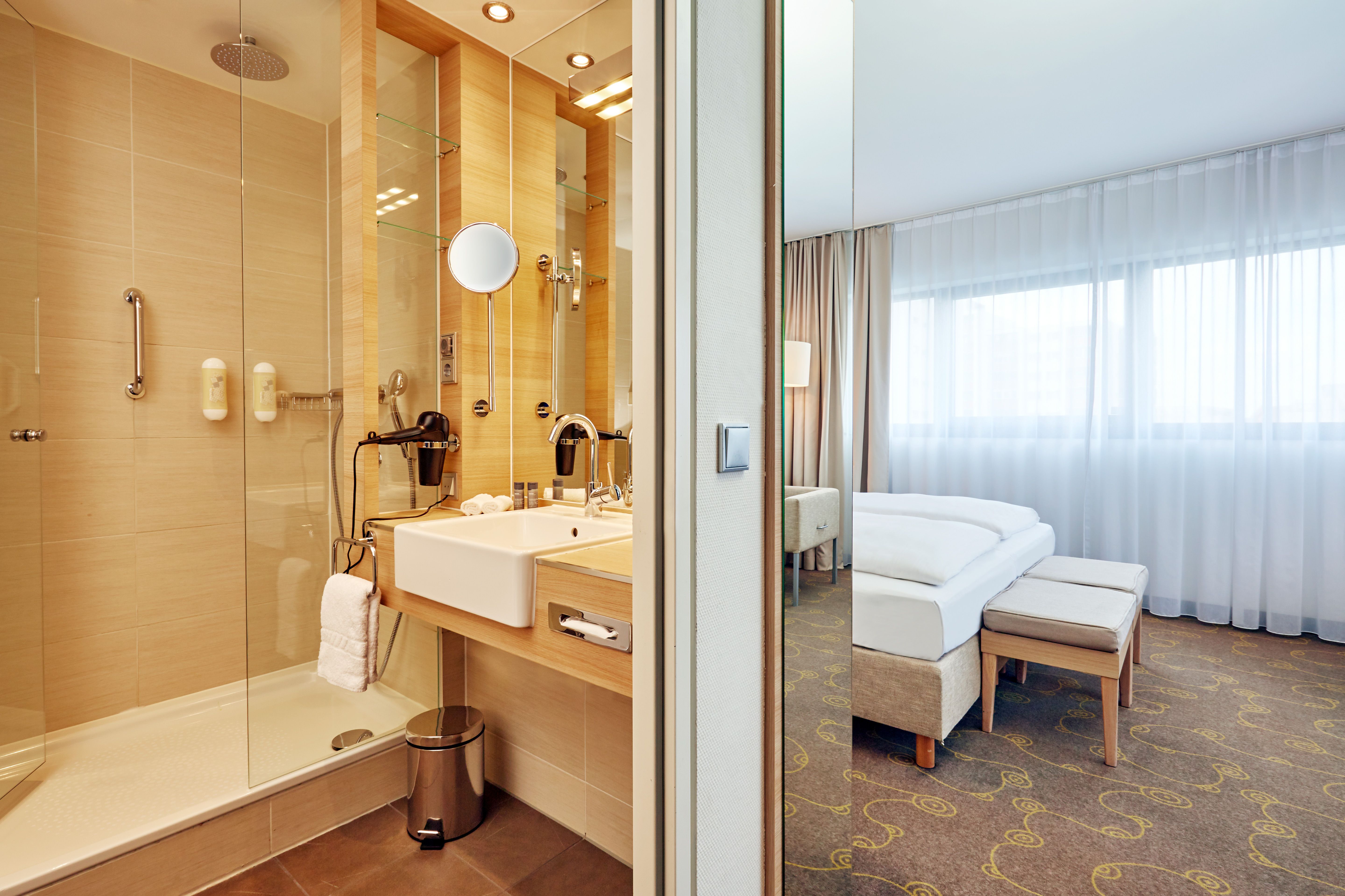 Modernes Hotelzimmer mit hell gestaltetem Bad, begehbarer Dusche und großzügigem Waschbecken. Heller, komfortabler Schlafbereich mit großem Fenster, Vorhängen und Doppelbett für erholsamen Aufenthalt.