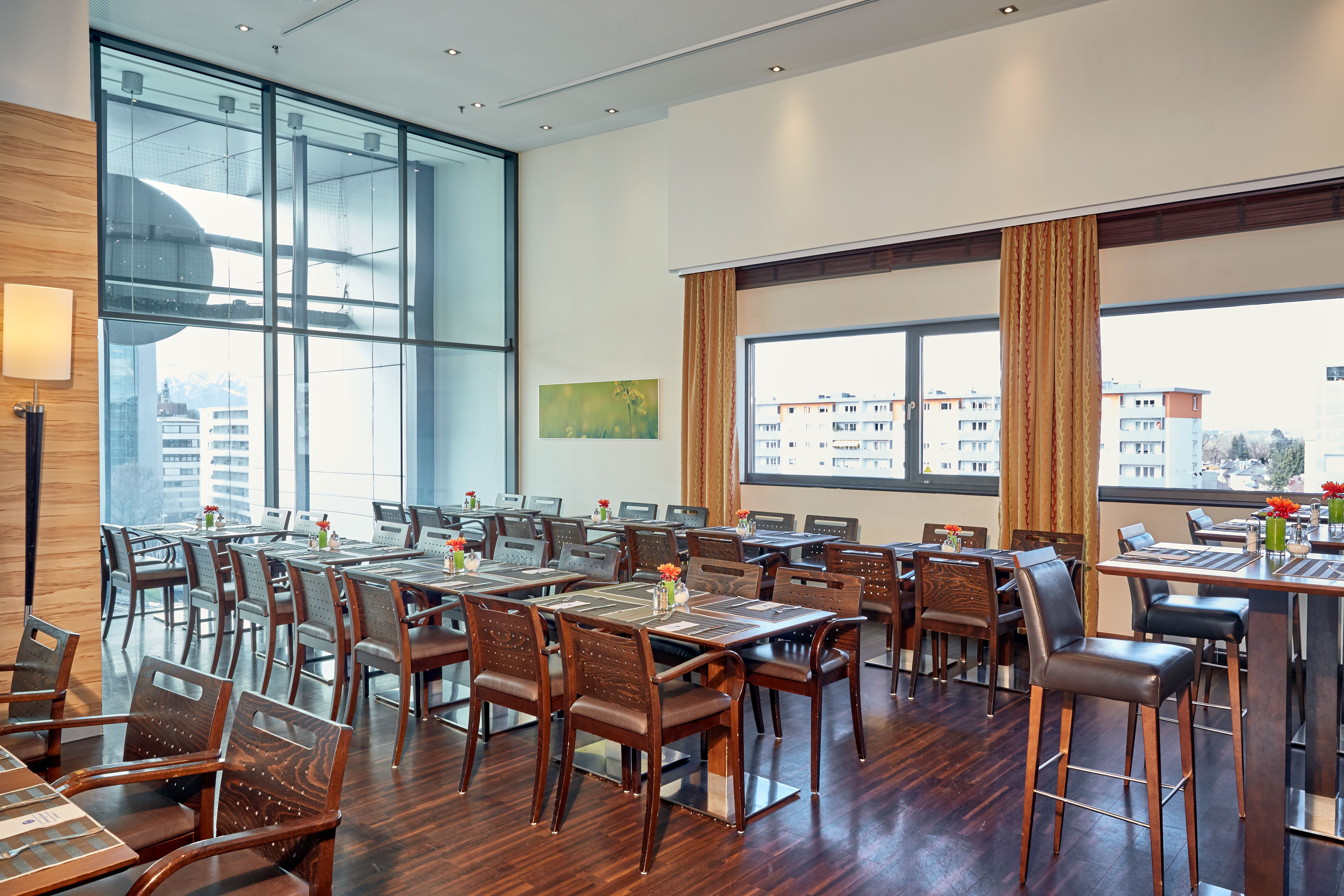 Moderner, heller Speisesaal mit Holzfußboden, großen Fenstern und zahlreichen Tischen, die mit Blumen dekoriert sind. Ruhige, einladende Atmosphäre für entspanntes Essen mit Ausblick auf die Stadt.
