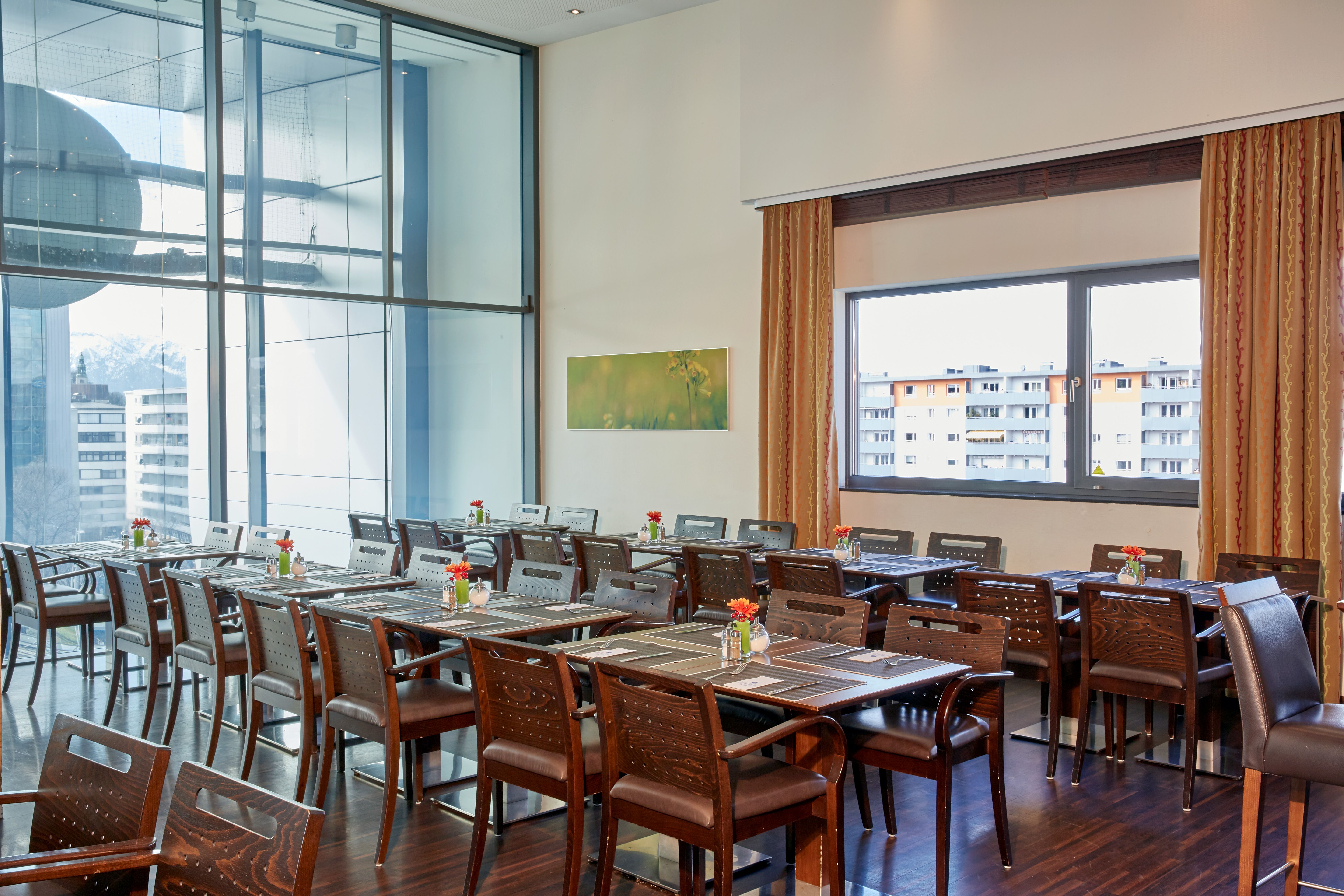 Helles, modernes Restaurant mit großen Fenstern, vielen Holztischen und Stühlen in Reihen, dekoriert mit kleinen Blumenvasen. Blick auf Stadtgebäude durch Fenster. Ruhige und einladende Atmosphäre für entspanntes Speisen.