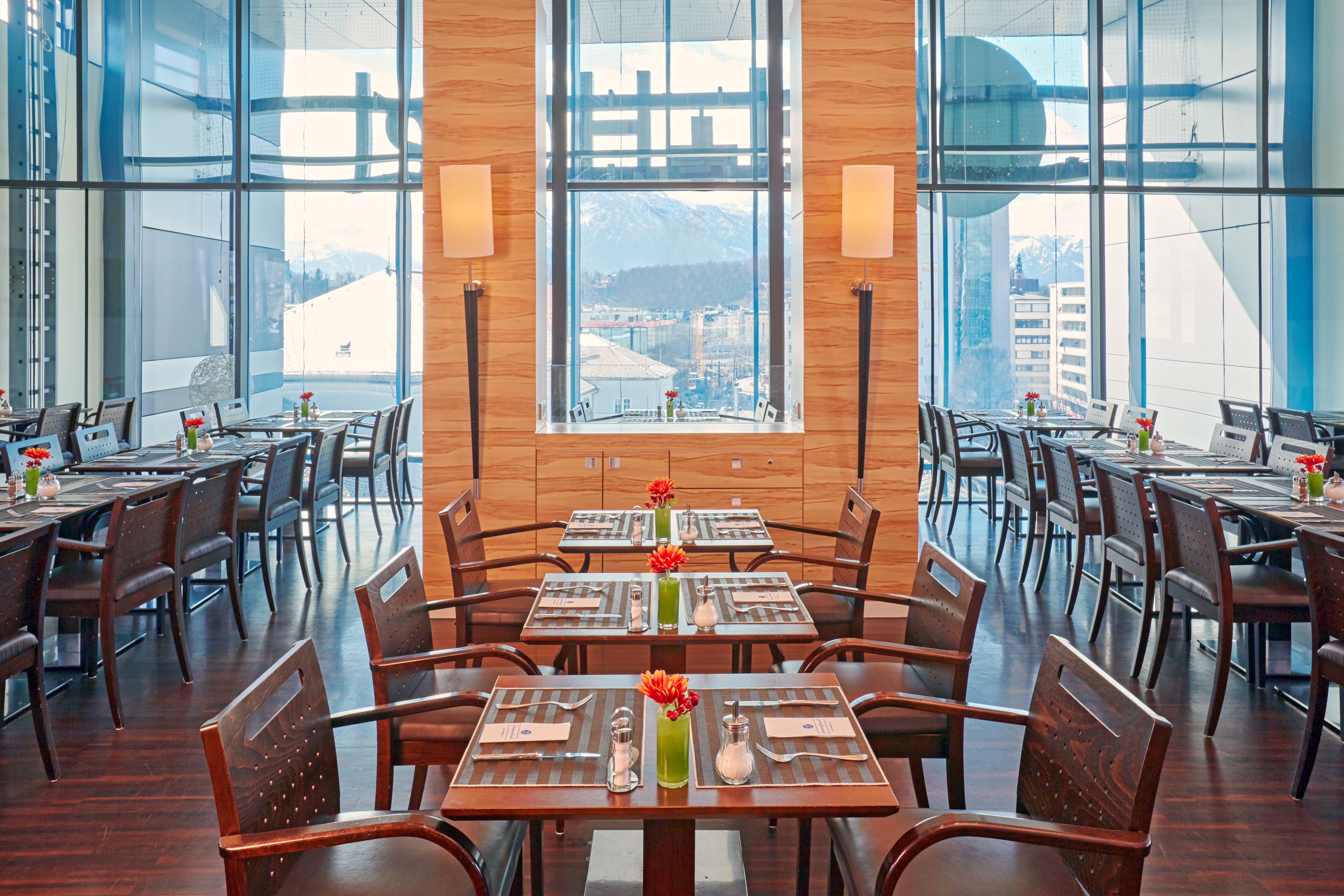 Modernes Hotelrestaurant mit großen Fensterfronten, hellem Holzakzent an der Wand und mehreren gedeckten Tischen mit Blumen, das viel Tageslicht und Ausblick auf die Stadt bietet.