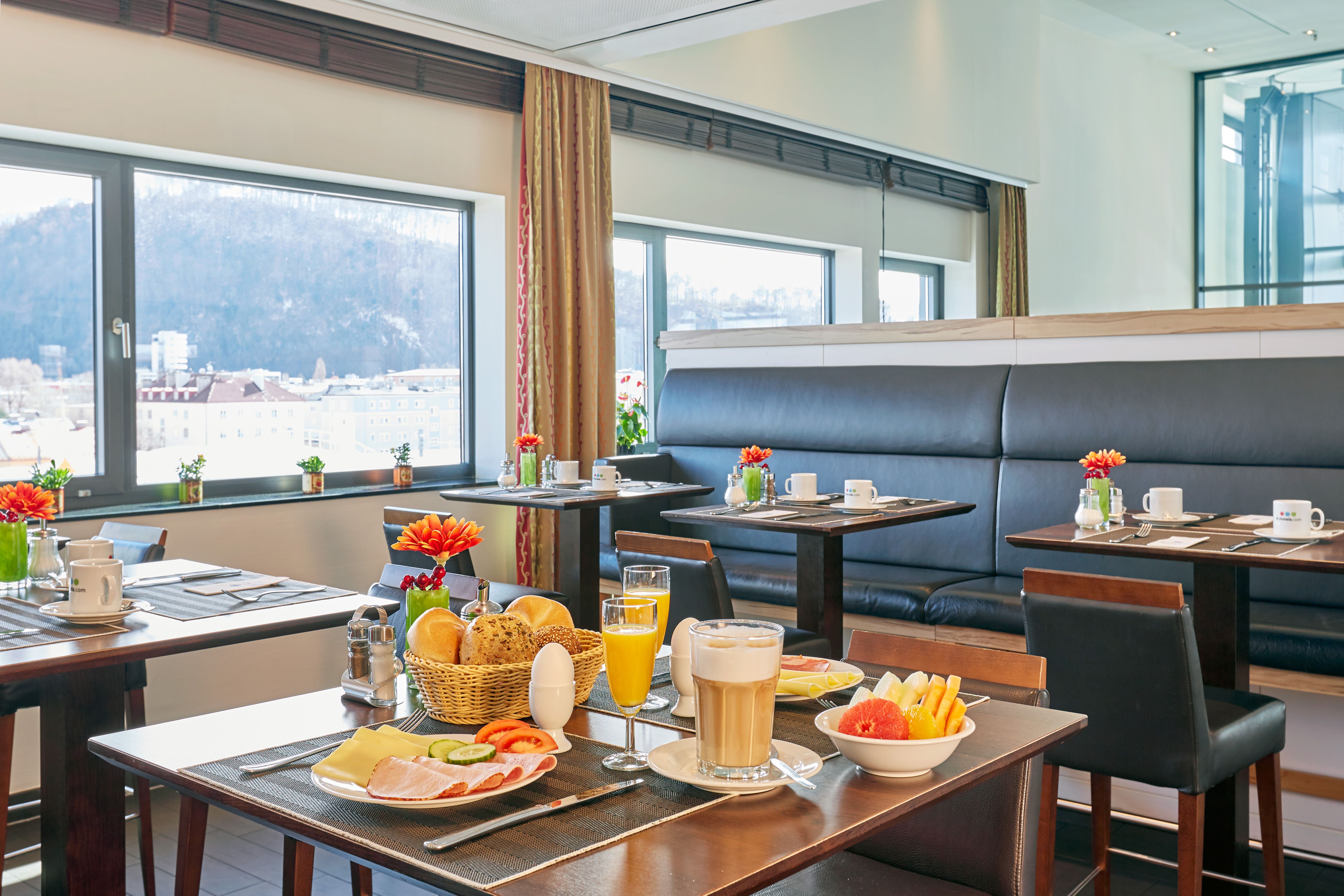 Helles, modernes Frühstücksrestaurant mit großen Fenstern und Bergblick. Tische sind gedeckt mit frischem Frühstück, darunter Kaffee, Orangensaft, Brot, Aufschnitt und Obst. Gemütliche Atmosphäre für entspannten Start in den Tag.